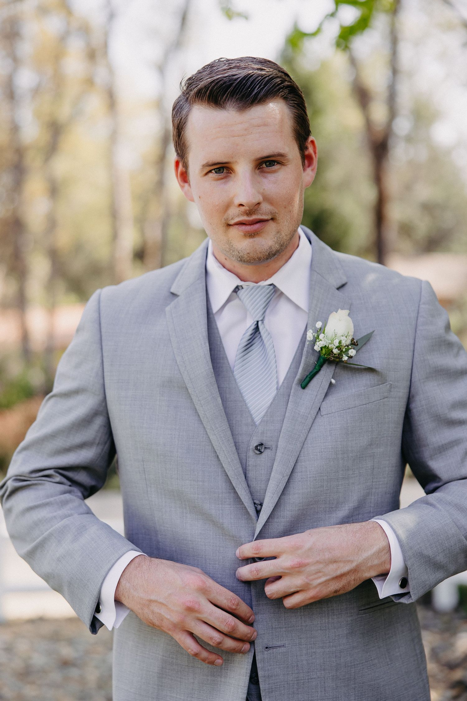 Close-up portrait showing a grey suit with silver tie and white boutonniere against autumn foliage.