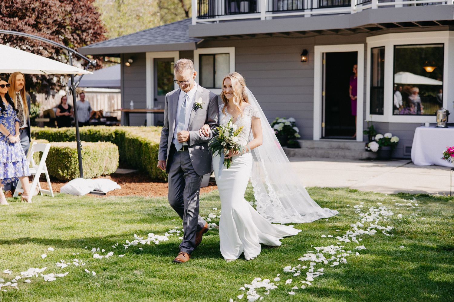 A wedding celebration on a sunny day at a grey house with flower petals scattered on the lawn.