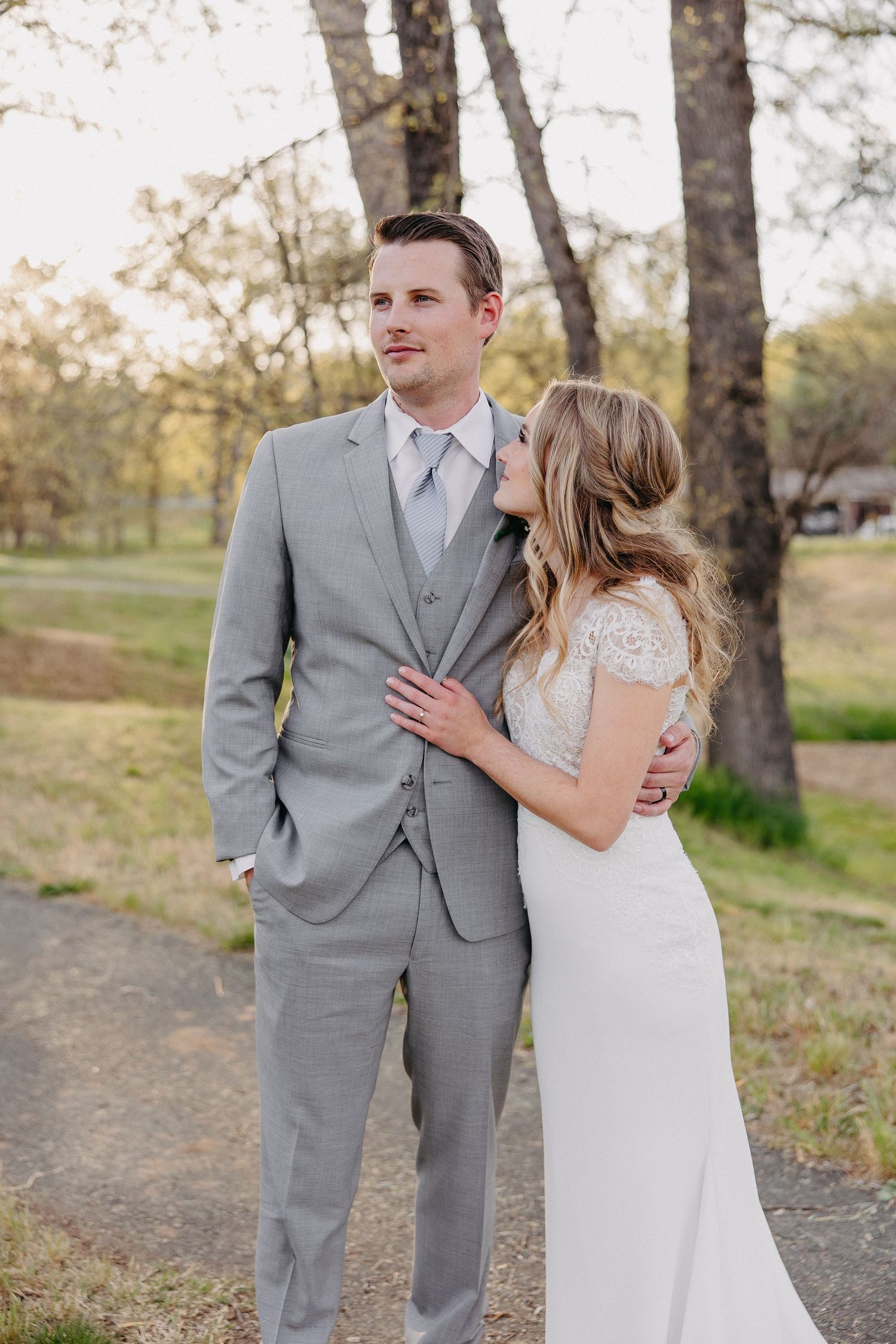 Elegant wedding couple in gray suit and white dress pose together on a natural path.