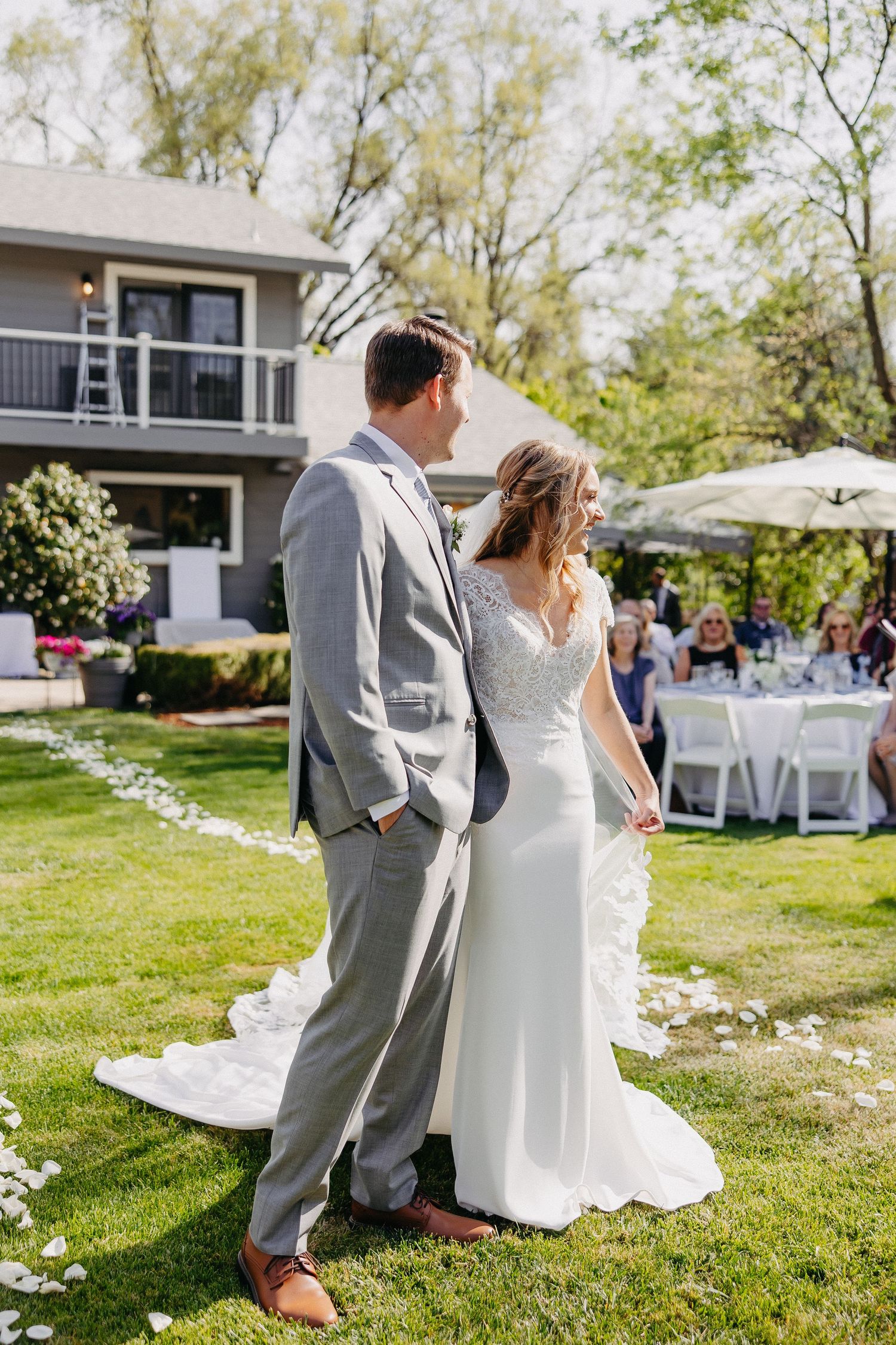 Newlyweds standing on lawn with reception setup and white folding chairs visible in background.