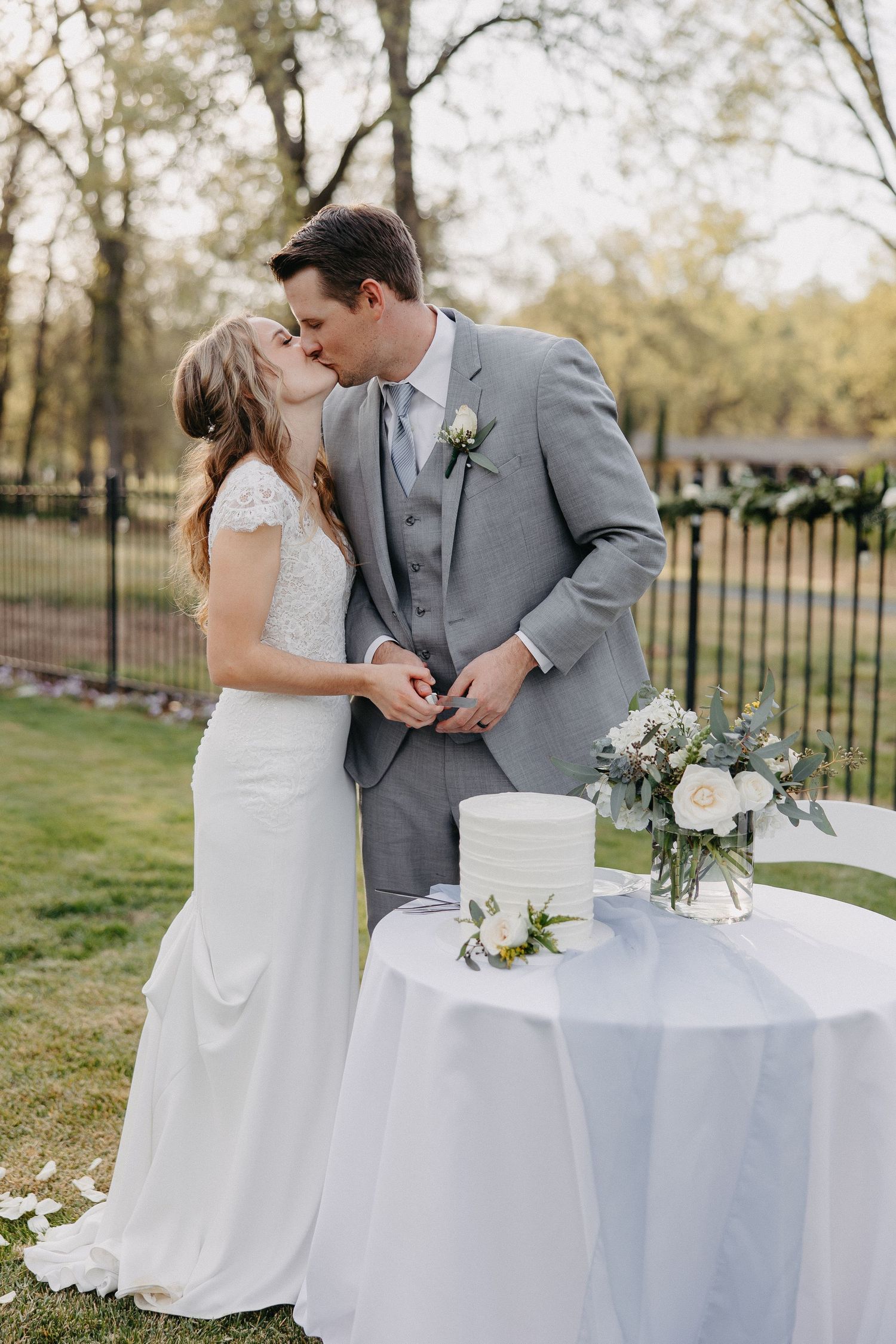 Romantic moment shared by couple in grey suit and white dress beside wedding cake outdoors.