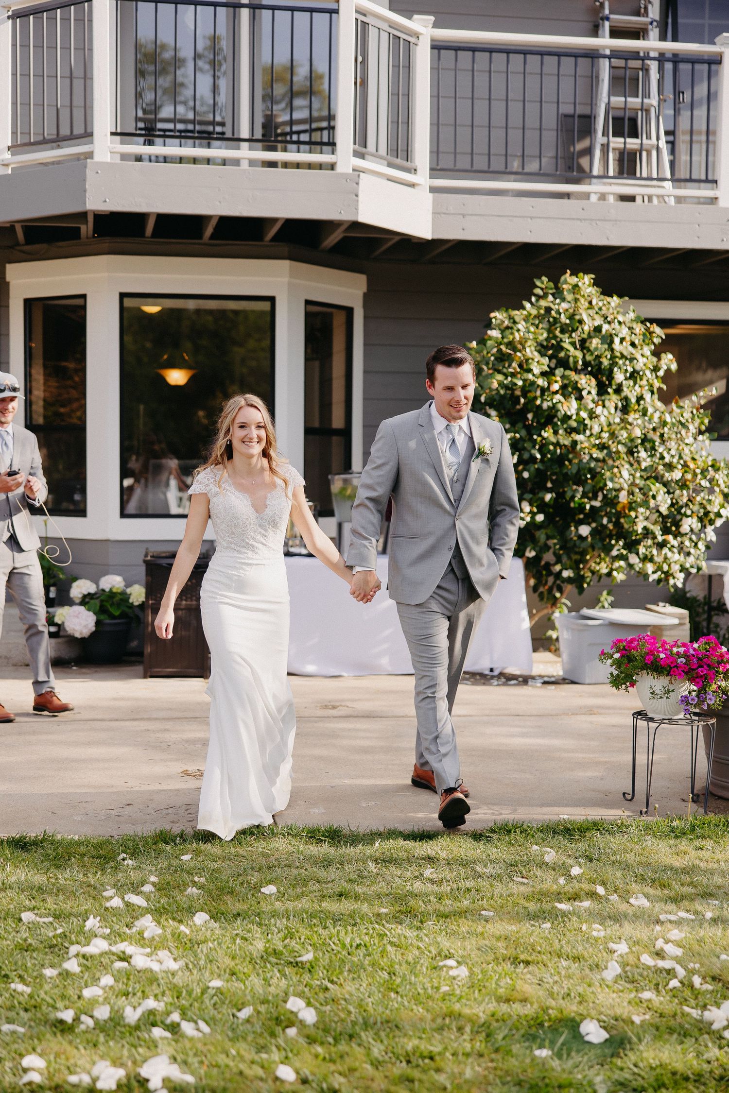 Newlyweds walking hand in hand on lawn with white flower petals scattered around.