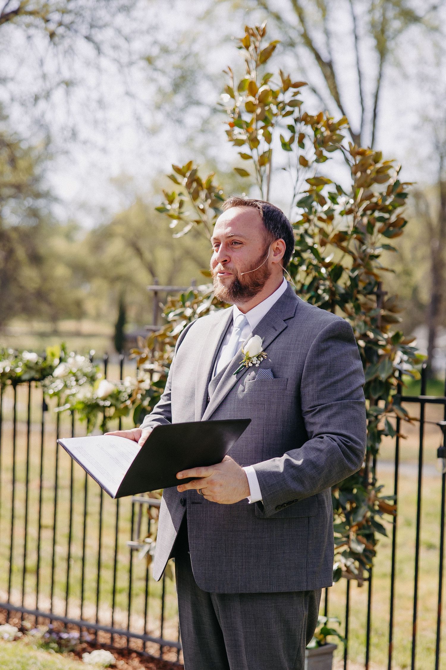 Wedding officiant in grey suit holding ceremony binder during outdoor service.