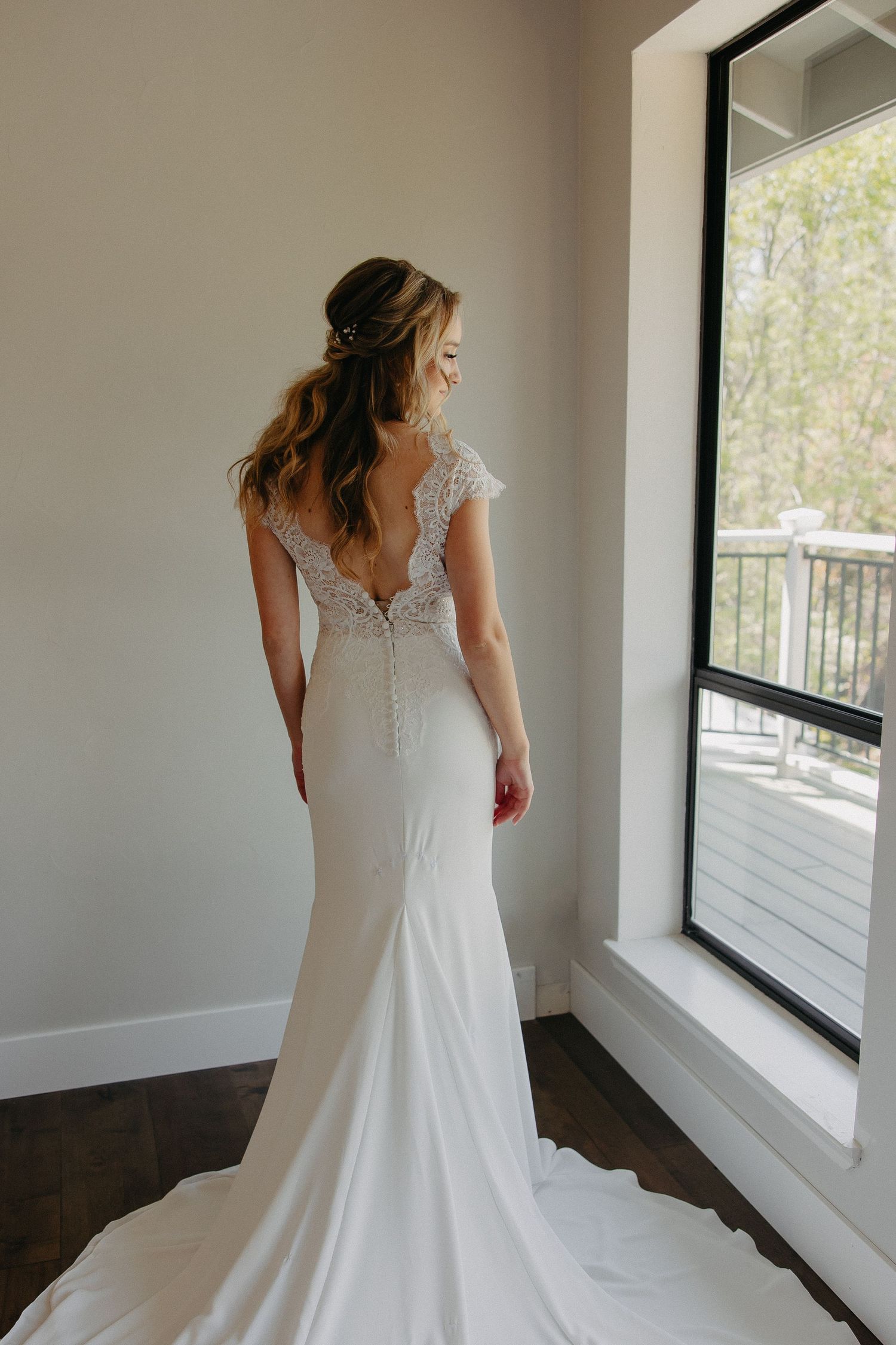 Elegant wedding dress with open back lace detail and flowing train by window.