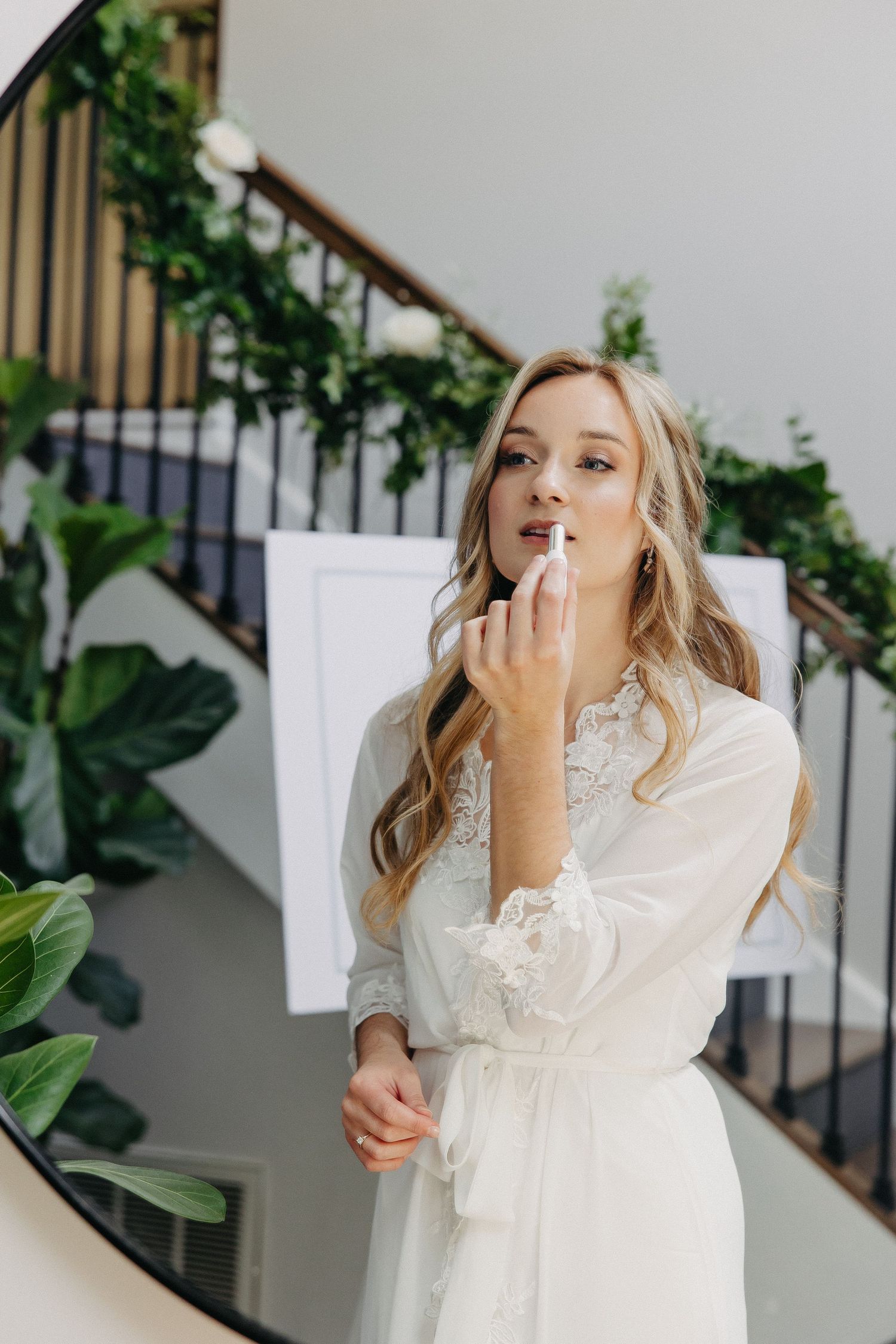 Elegant white lace robe being worn while applying makeup in front of a staircase with greenery.