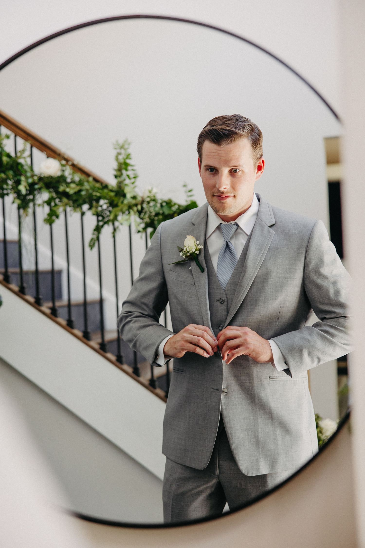 Mirror reflection capture of formal grey suit and boutonniere with curved staircase in background.