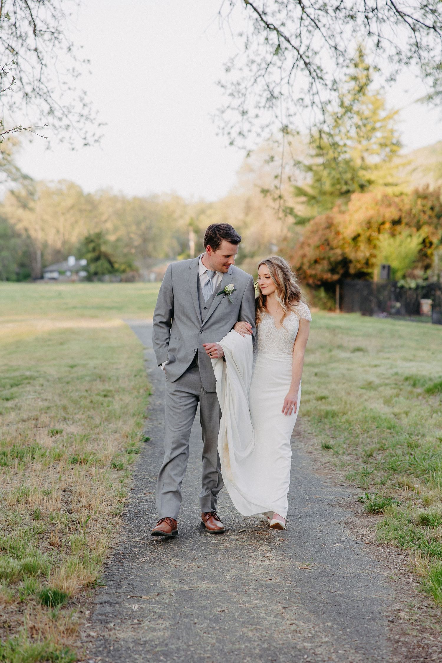A couple in wedding attire walking together and smiling on a tree-lined path during golden hour.