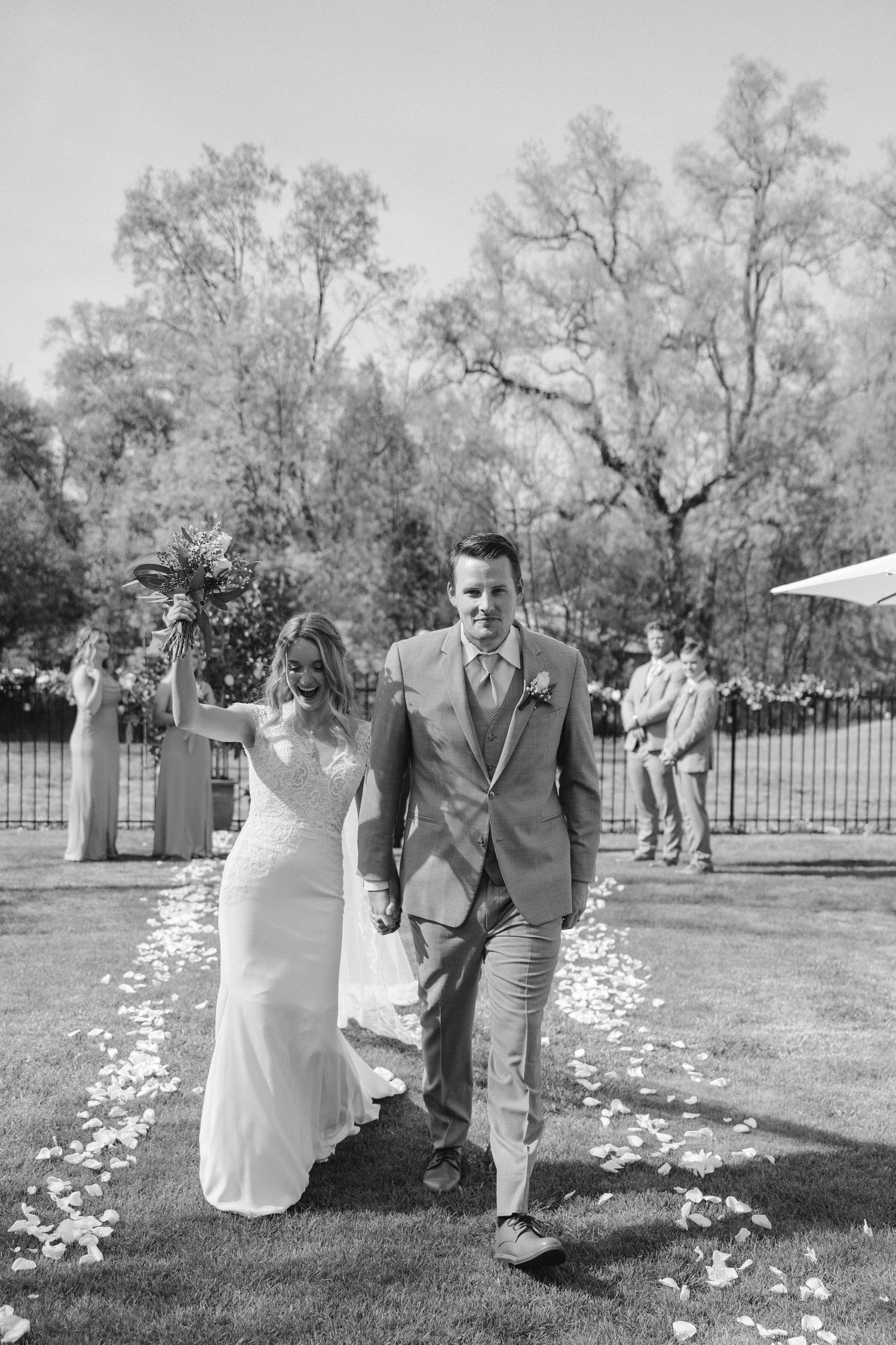 A newlywed couple walks down the aisle after their outdoor ceremony with flower petals scattered on the grass.