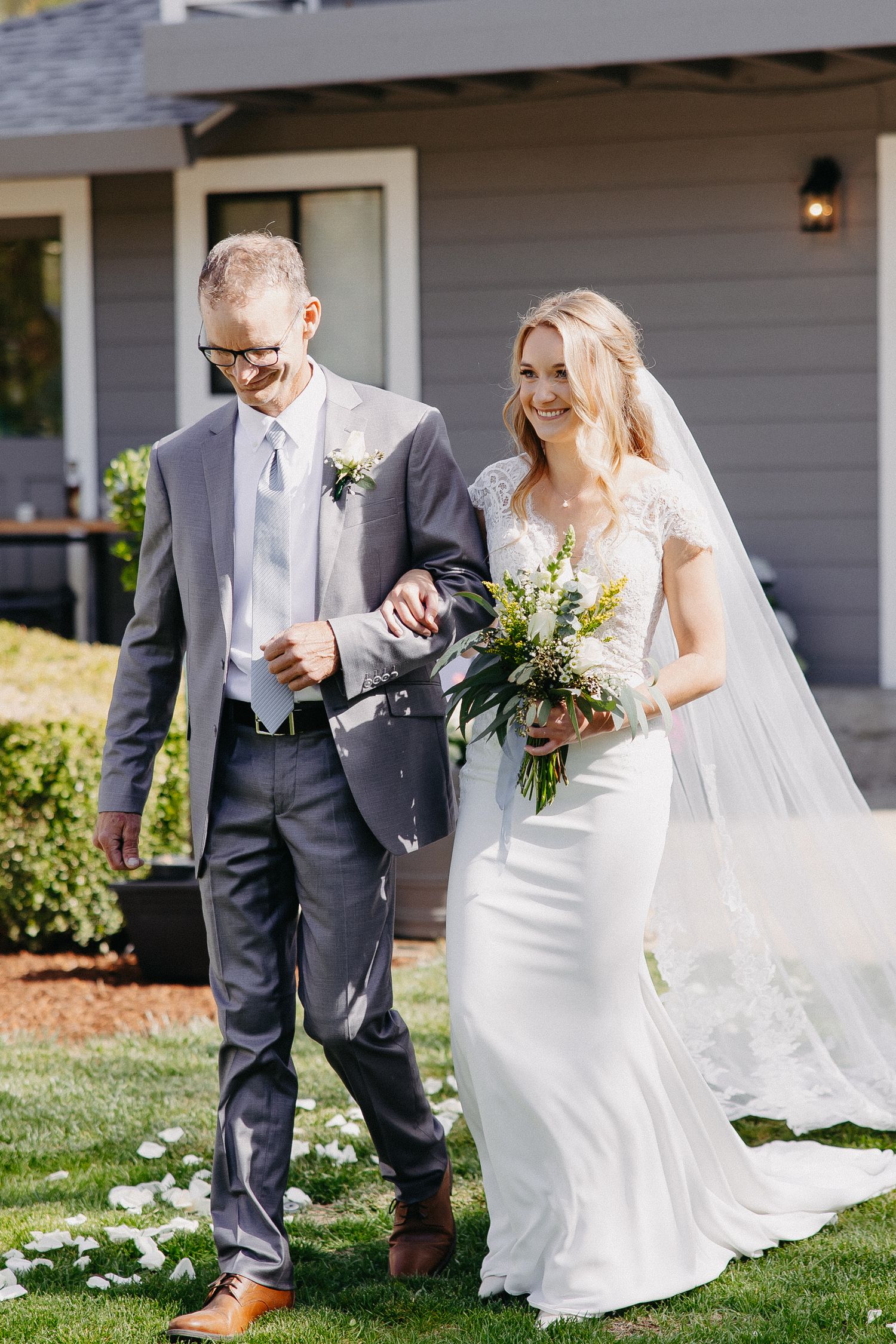A wedding couple walks together on green grass with scattered white petals outside a grey house.