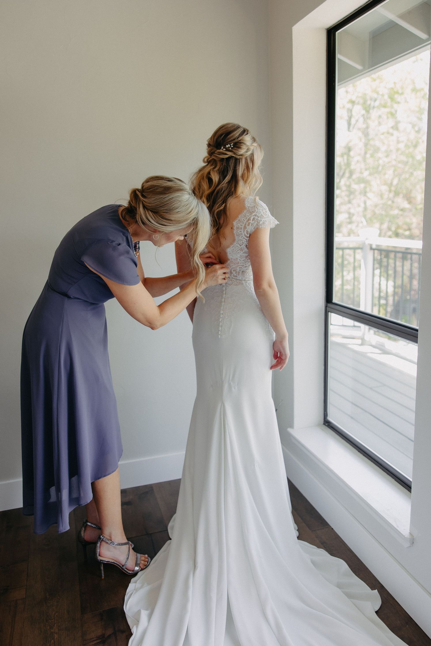 Bridesmaid in navy dress helping with final wedding gown adjustments near a bright window.