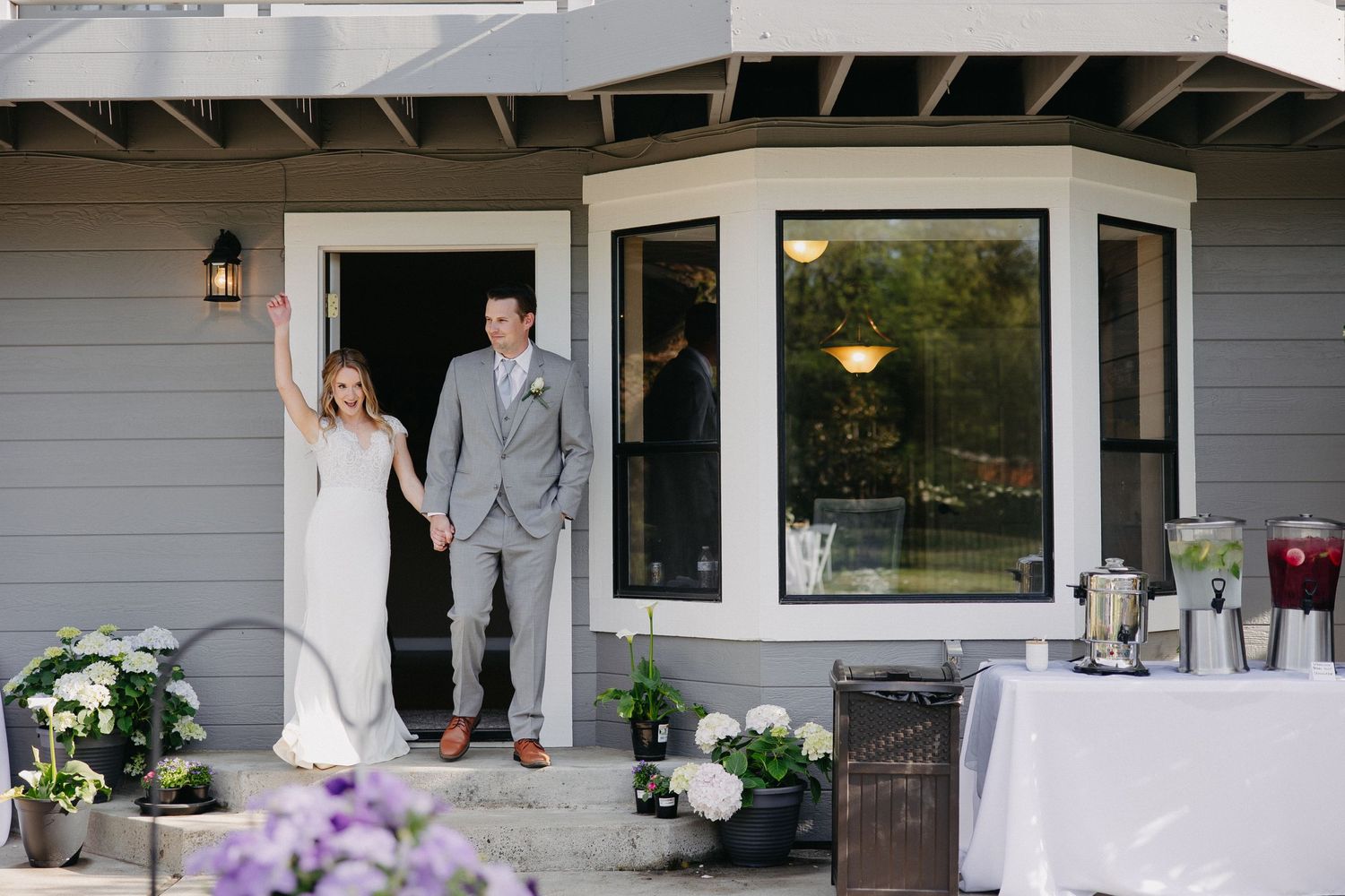 Wedding couple stands on porch with reception setup and purple flowers in foreground.