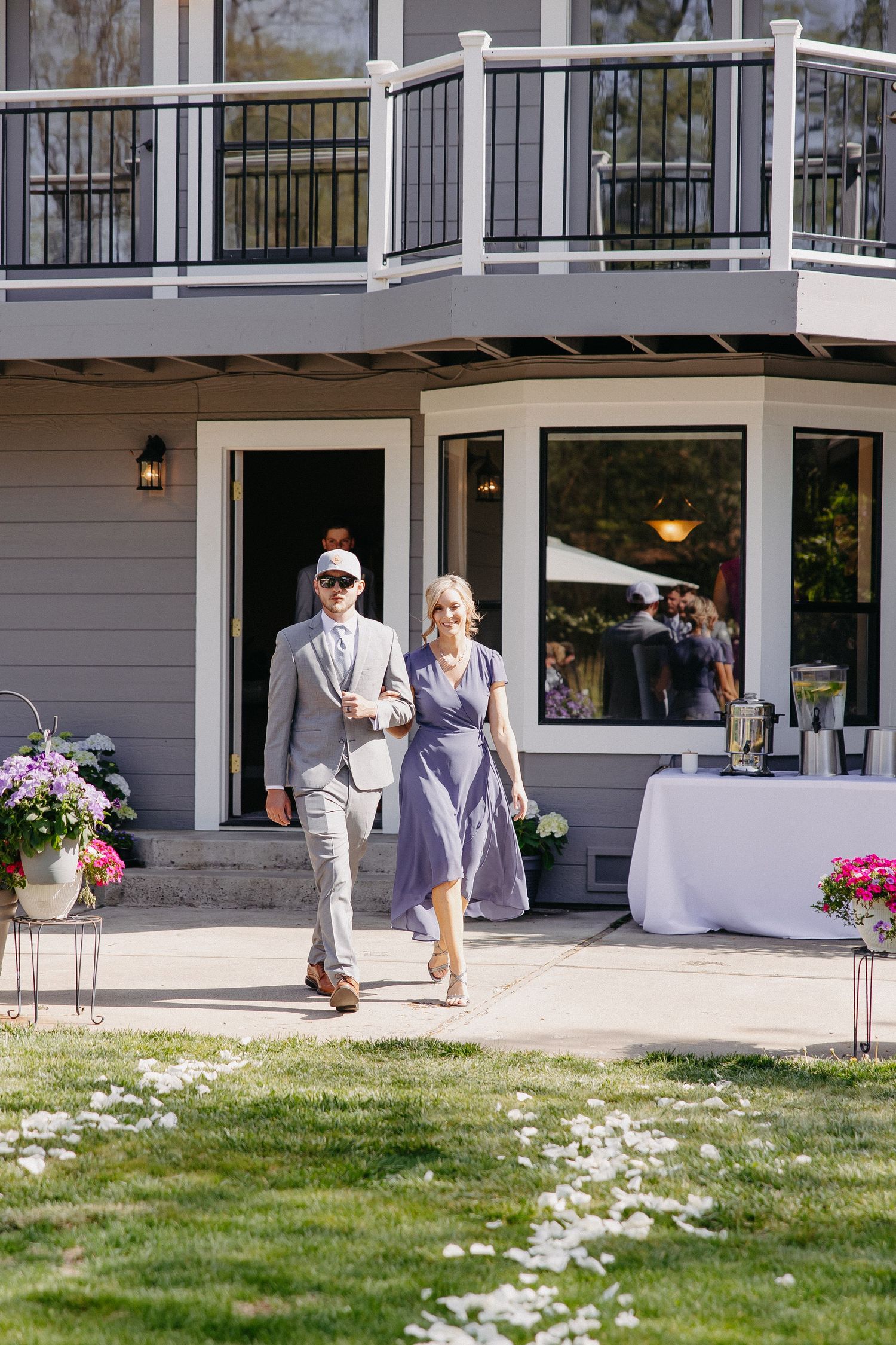 A couple walks across a lawn decorated with flower petals and outdoor reception tables.