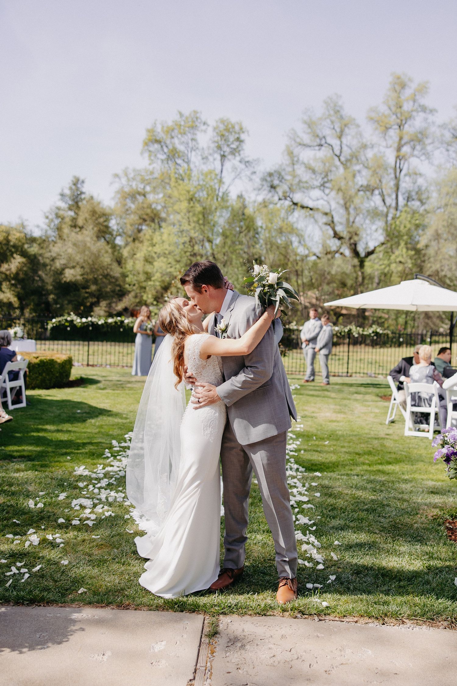 A couple shares their first kiss as newlyweds on a sunny lawn with scattered flower petals.