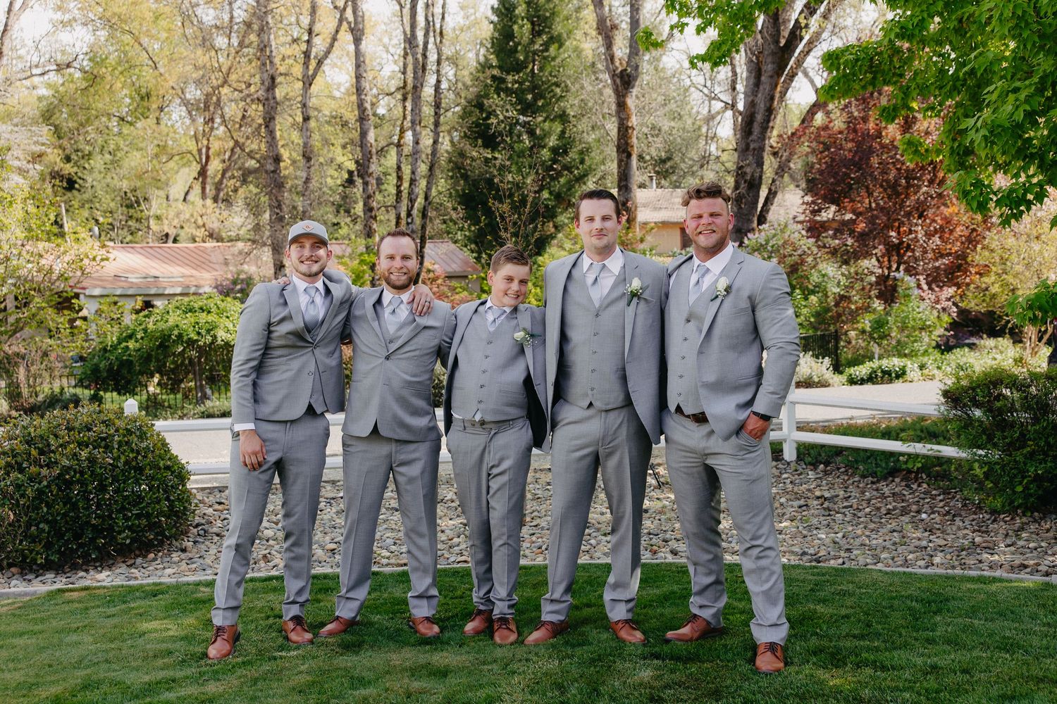 Groomsmen in matching gray suits and brown shoes pose together in a garden setting.