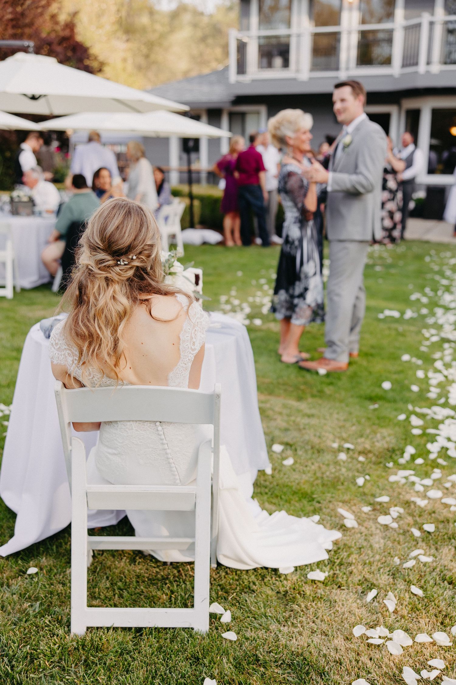 Elegant outdoor wedding reception with bride seated at table while guests mingle on lawn with scattered flower petals.