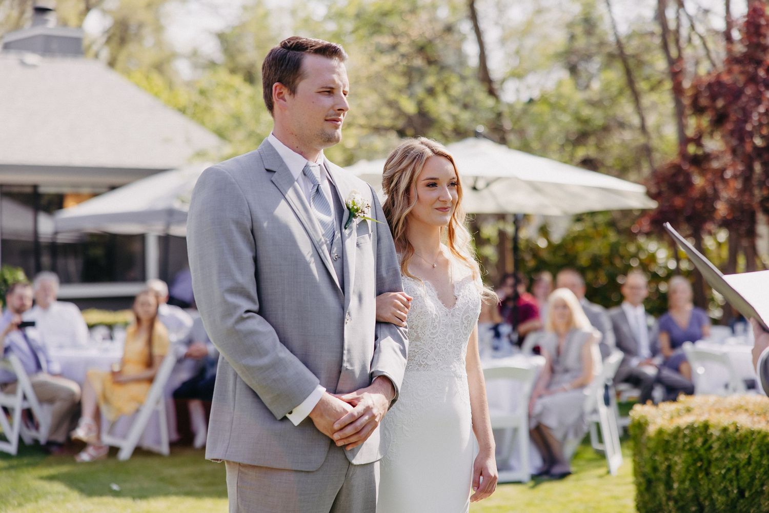 A bride and groom stand together at an outdoor garden wedding ceremony as guests look on.