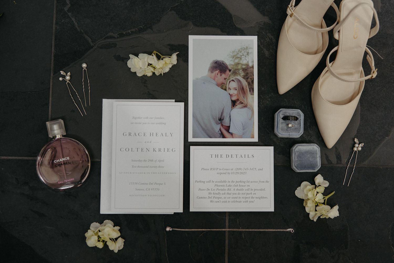 Wedding invitation suite with elegant stationery, beige shoes, perfume bottle and floral accents on dark surface.