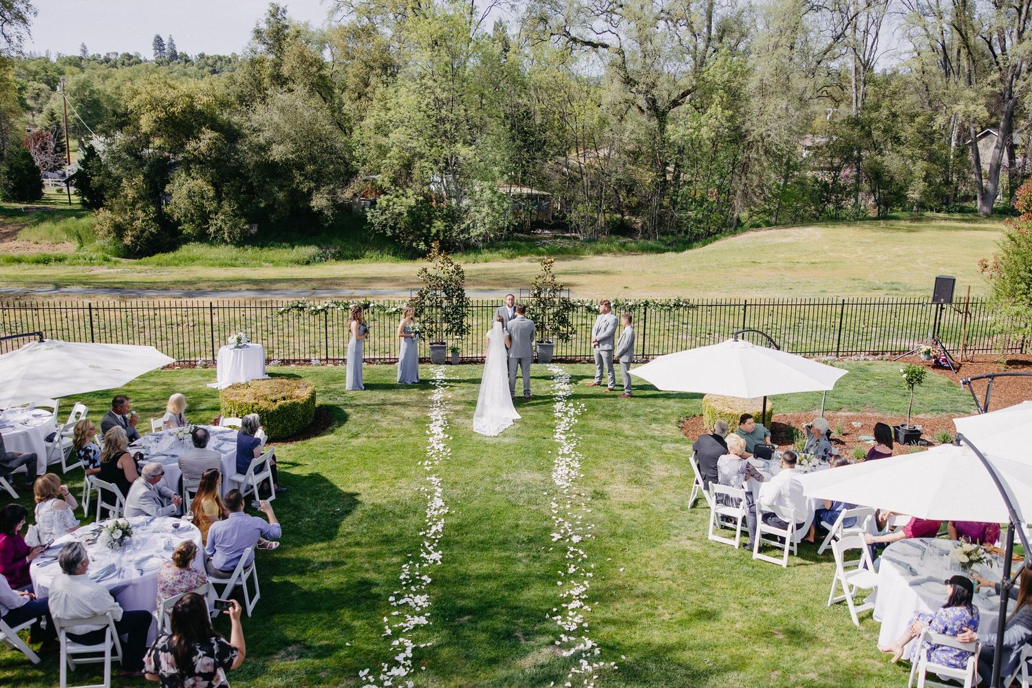 Outdoor garden wedding ceremony with white chairs and petal-lined aisle under blue sky.