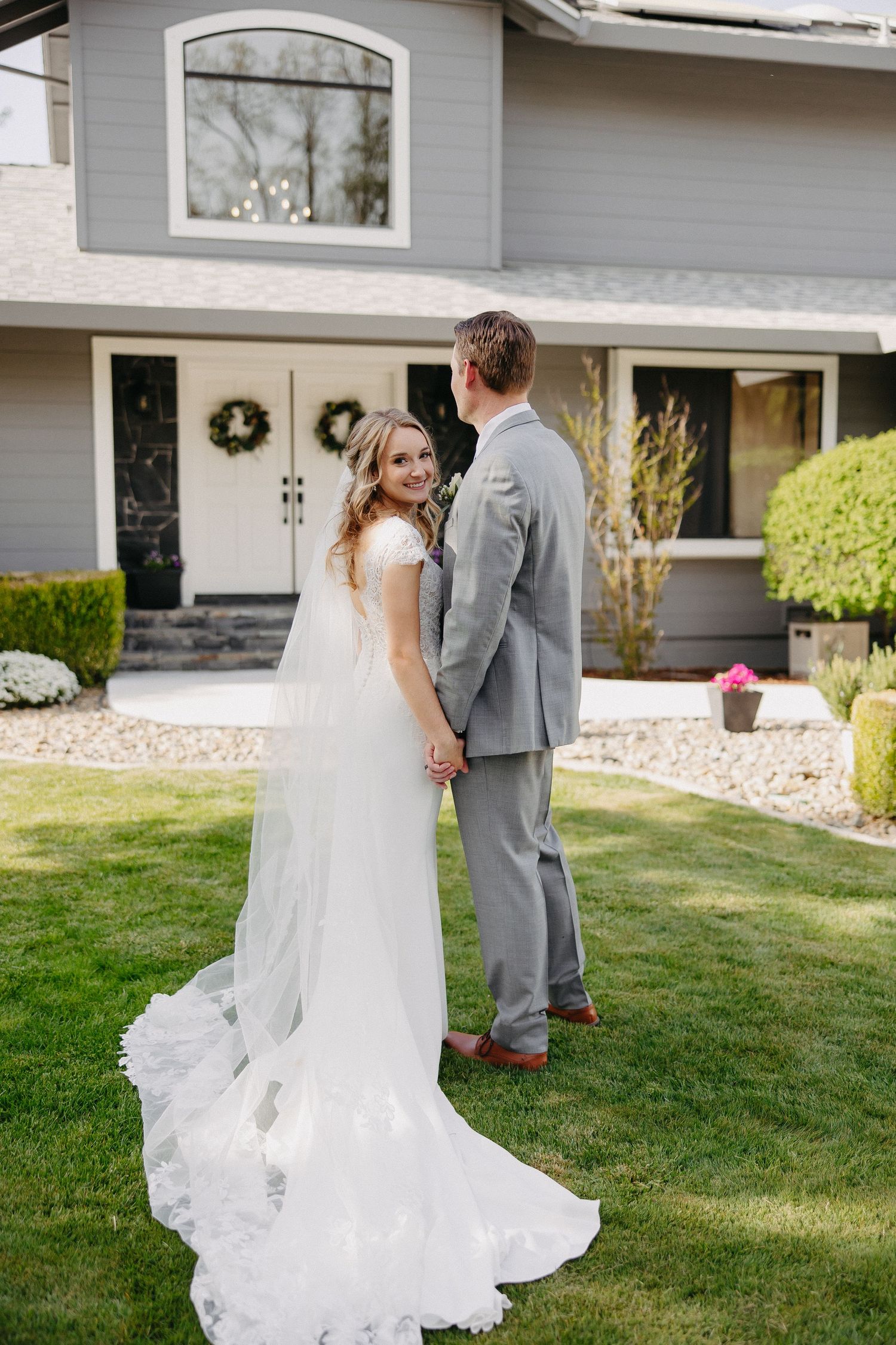 Elegant couple in wedding attire pose together on a landscaped lawn with house backdrop.