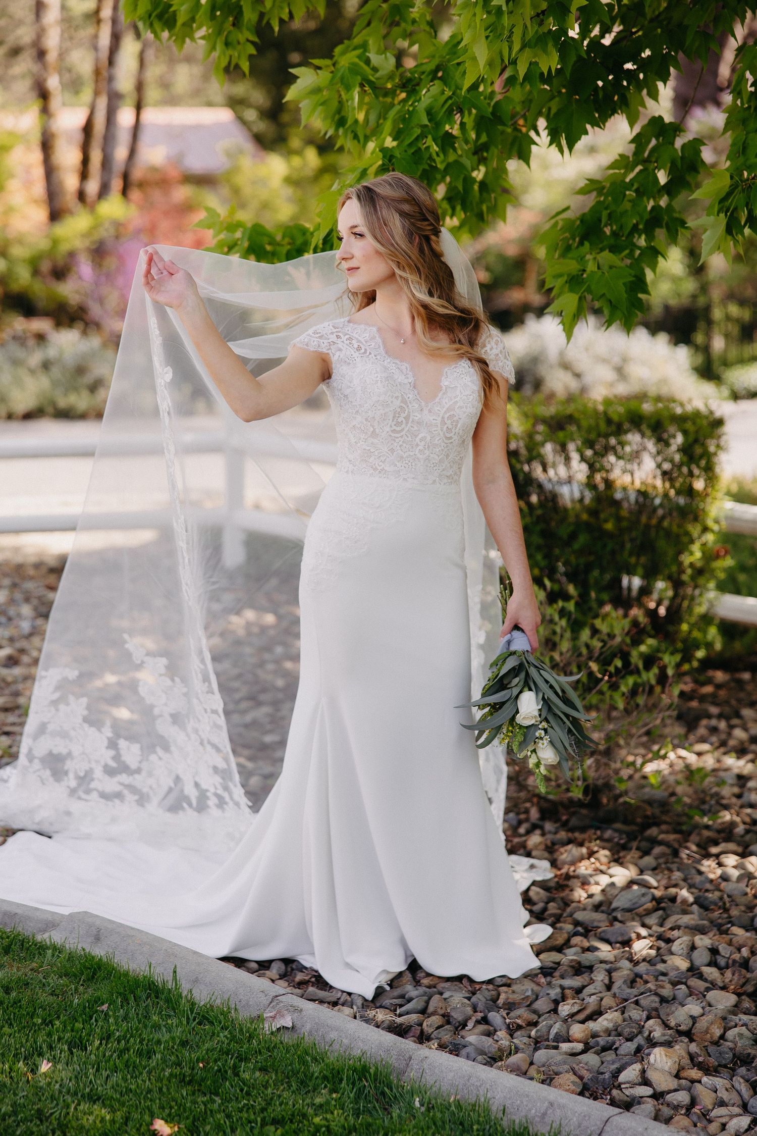 Bride adjusting veil in garden setting while holding eucalyptus bouquet.