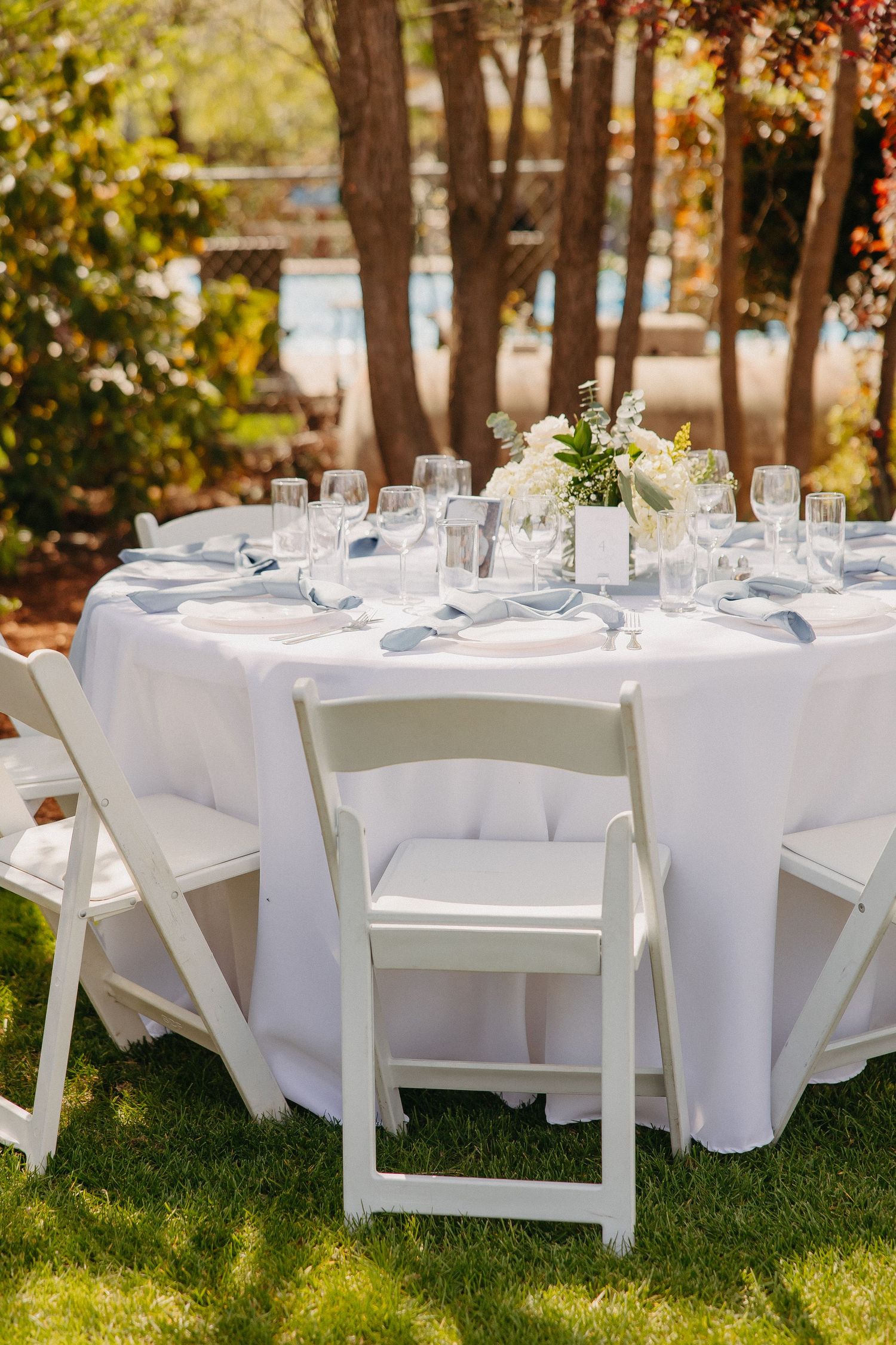 Elegant outdoor table setting with white linens and folding chairs in natural sunlight.