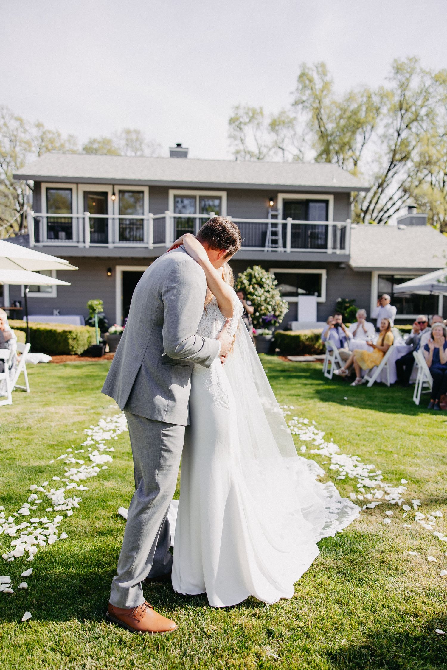 A romantic wedding moment captured on a lawn with scattered petals and grey house backdrop.