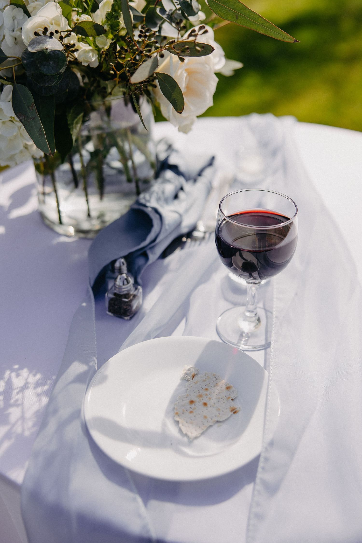 A glass of red wine and floral centerpiece on white tablecloth in sunlight.