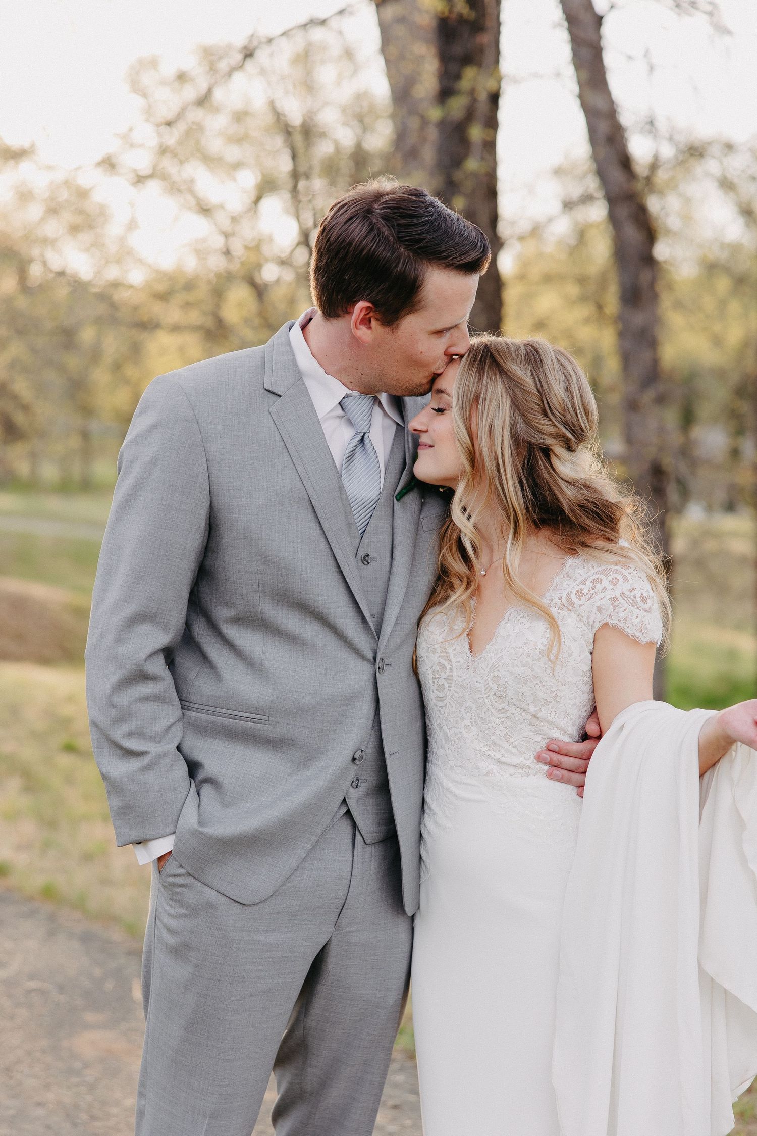 Intimate moment between newlyweds in grey suit and lace dress outdoors.