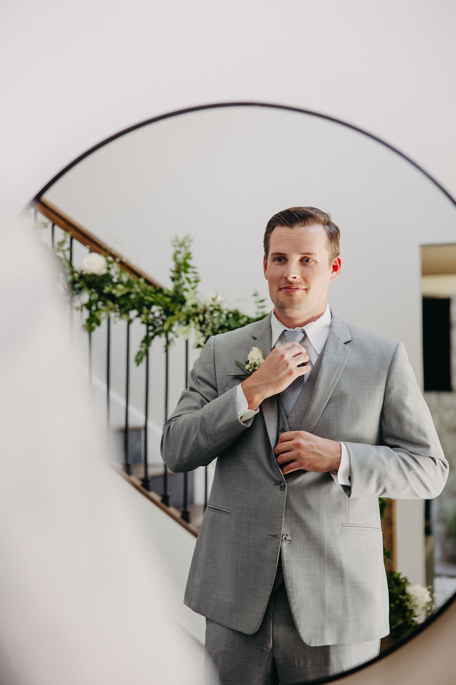 Elegant mirror shot showing grey suit adjustments with decorative staircase and greenery.