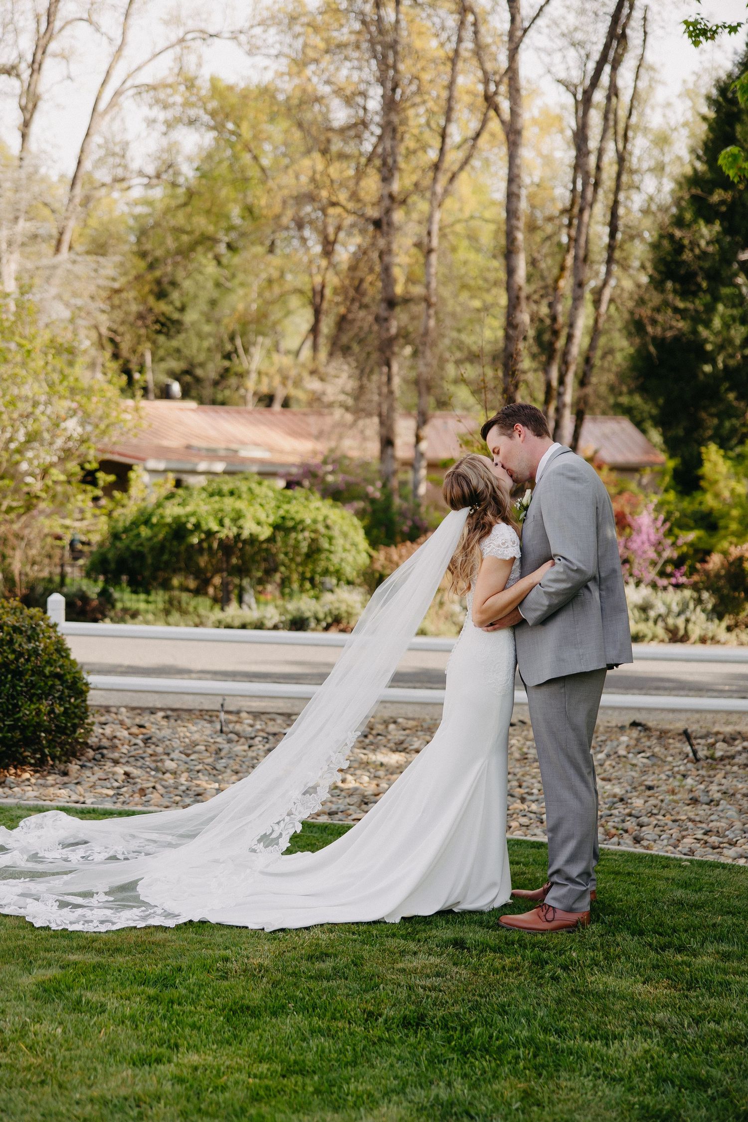 A romantic wedding couple sharing a kiss outdoors with the bride's long flowing veil trailing behind them.