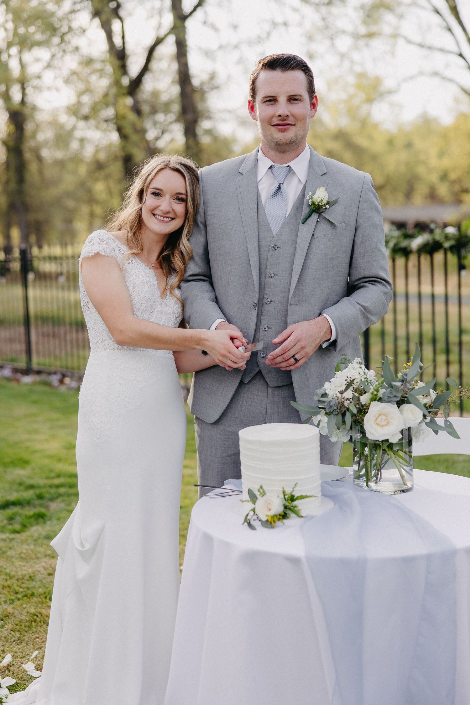 Elegant outdoor wedding scene with couple standing by white cake and floral arrangements on round table at garden venue.