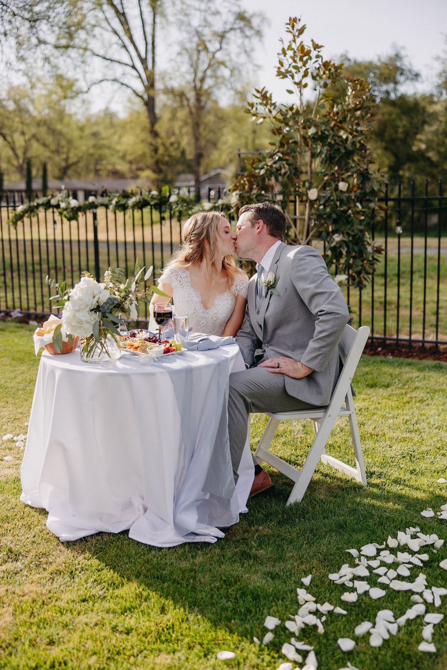 Romantic newlyweds share intimate moment at outdoor reception table decorated with white linens and flowers.
