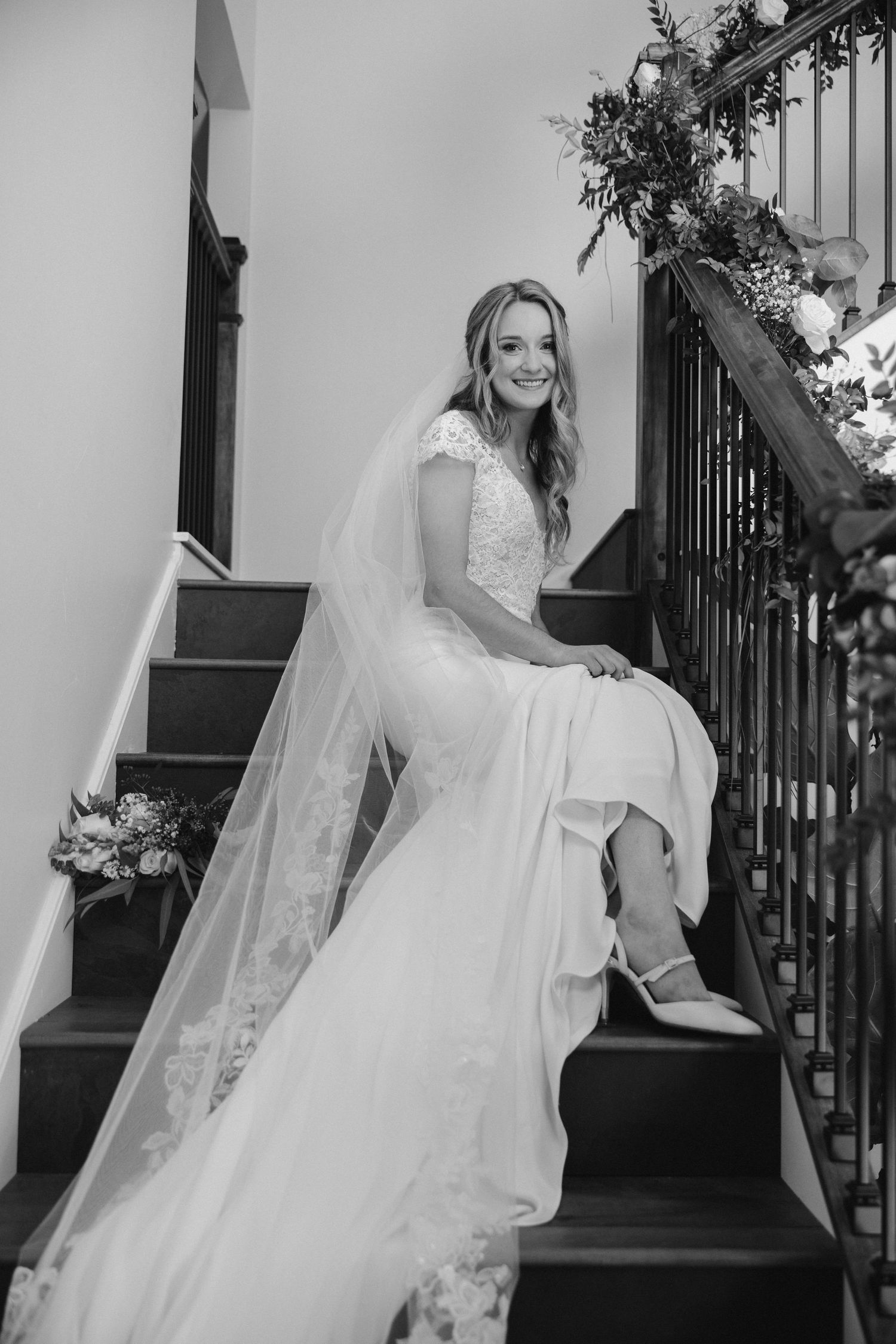 Black and white portrait of bride sitting on staircase decorated with floral arrangements.