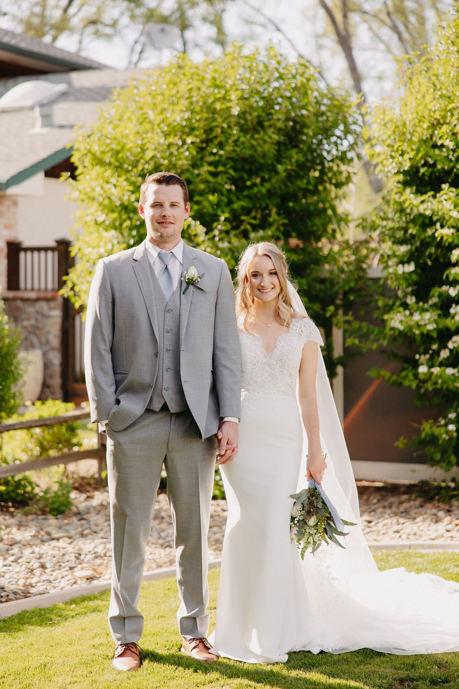 A full-length wedding portrait of a couple on a manicured lawn with greenery and architecture in background.