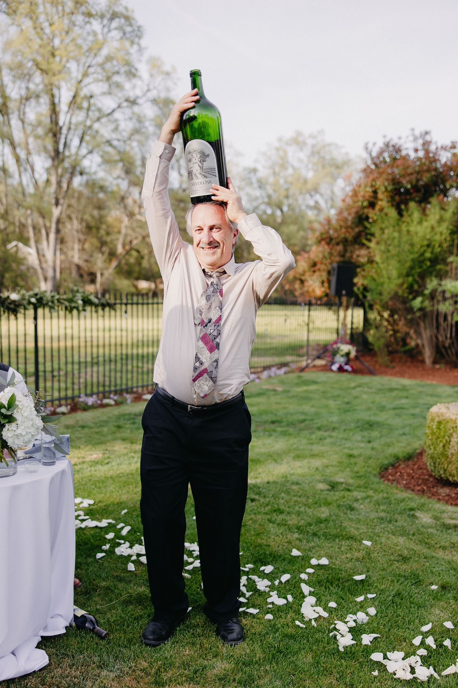 Wedding guest in tie celebrating with champagne bottle raised high in garden setting.