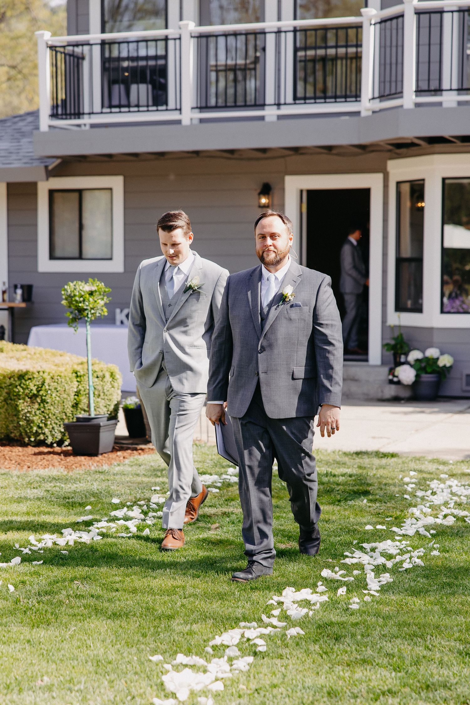 Two people in grey suits walk along a petal-strewn path during a wedding.
