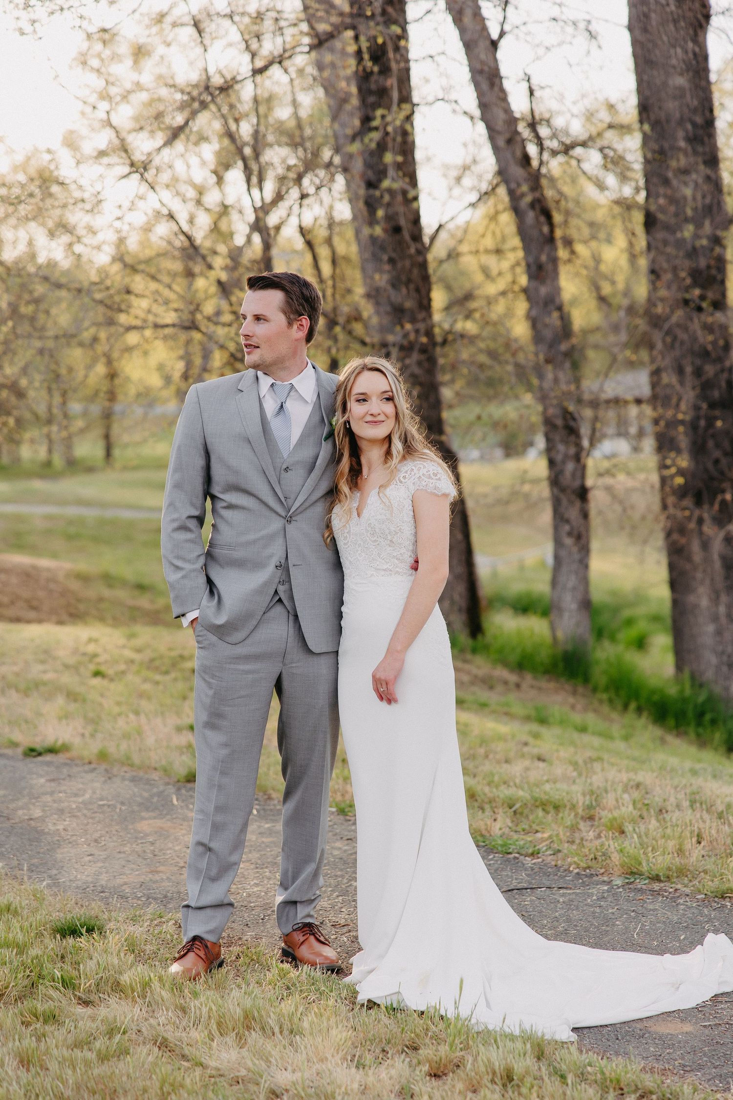 A wedding couple poses outdoors in formal attire against a backdrop of tall trees on a dirt path.