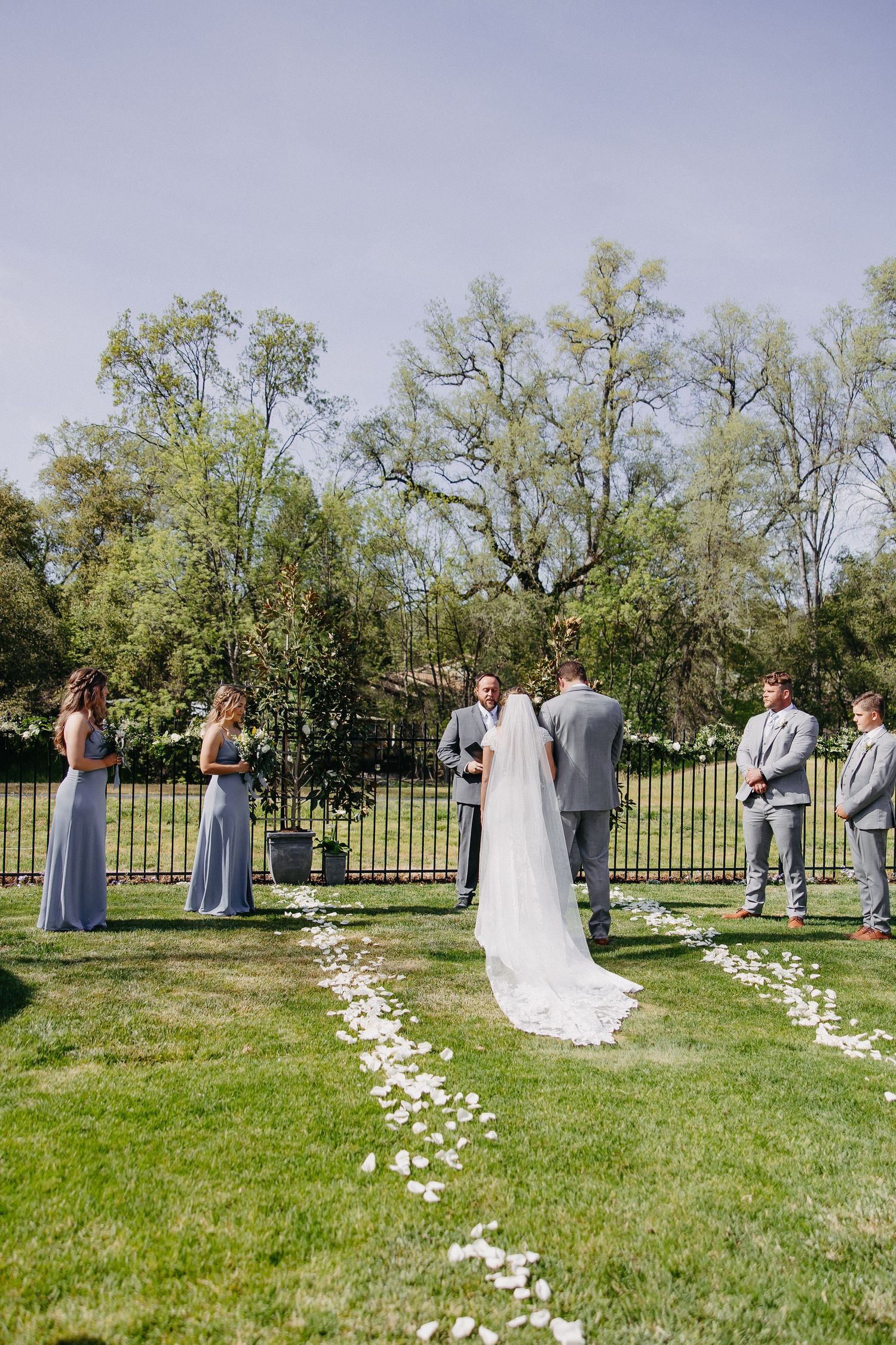 Outdoor wedding ceremony setup with petal aisle and iron fence in natural setting.