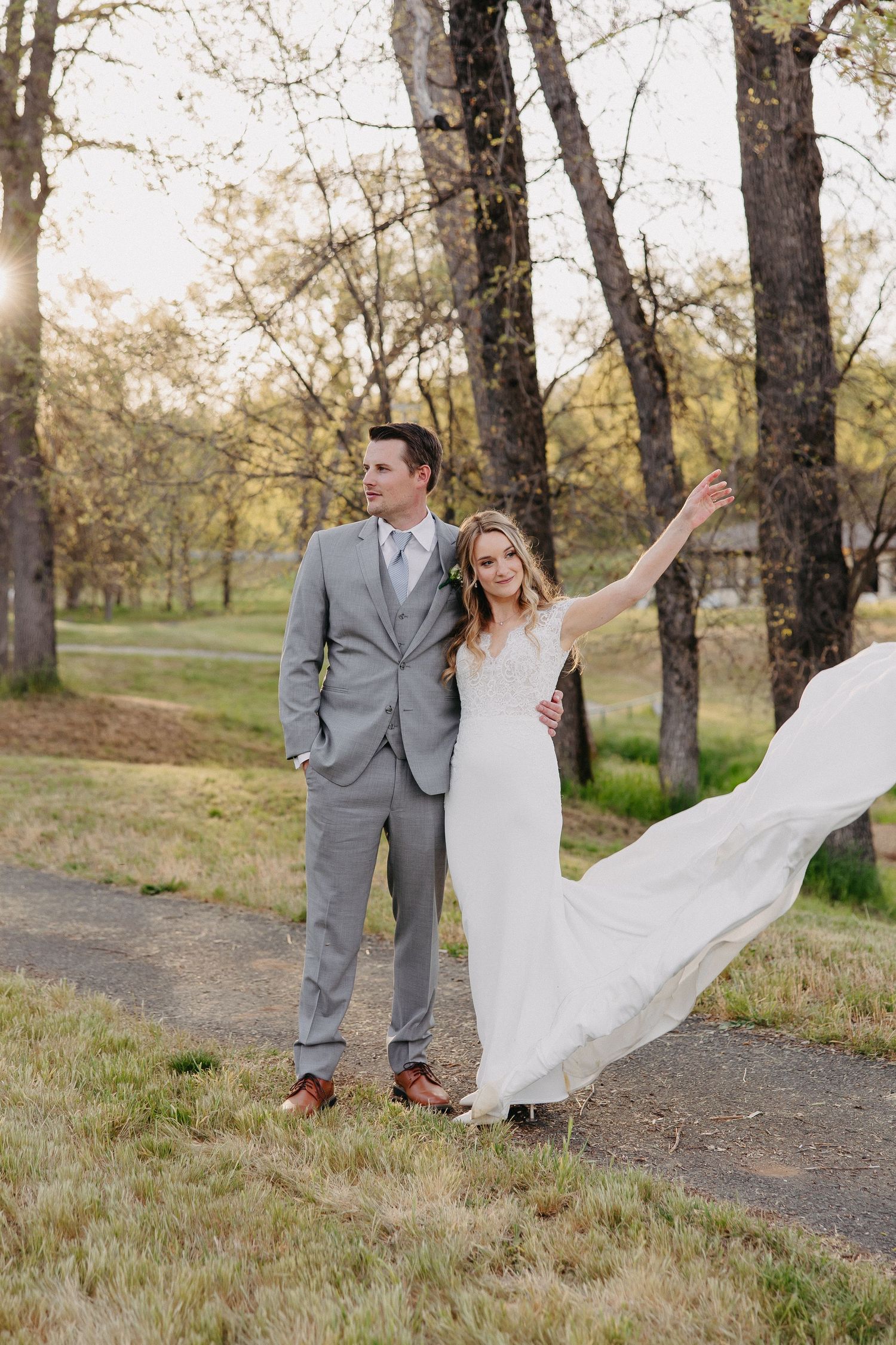 A bride in white dress twirls while standing next to groom in grey suit on nature path.
