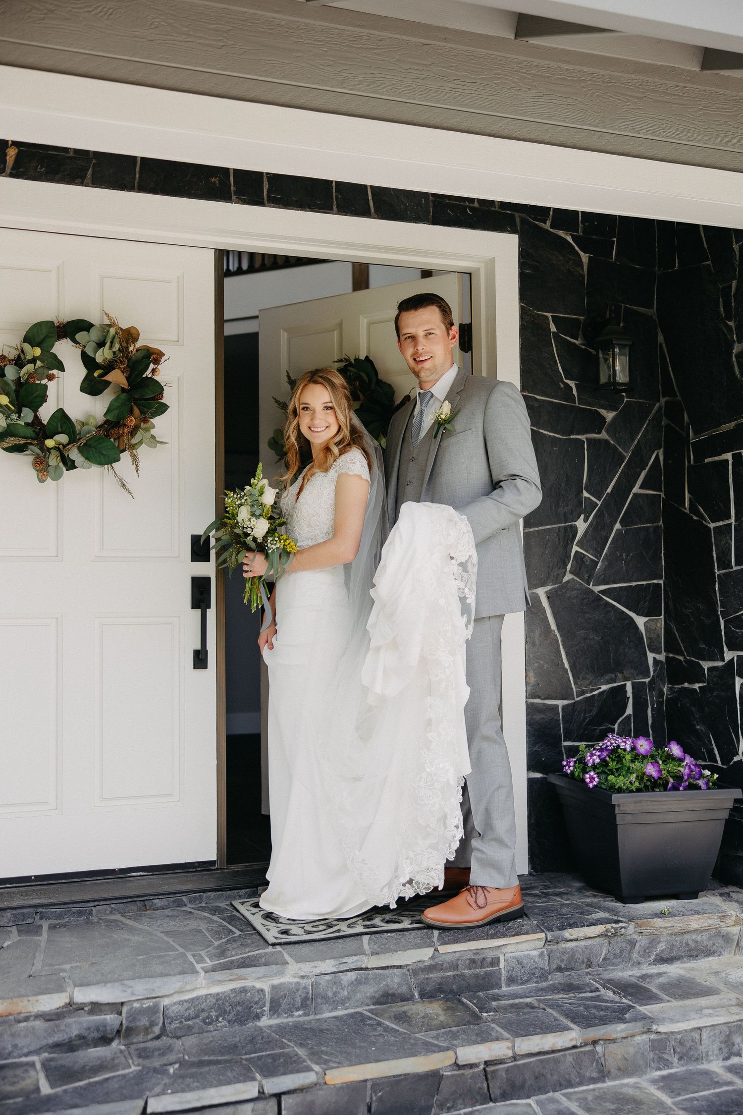Wedding couple poses in entryway with stone steps and decorative wreath on door.