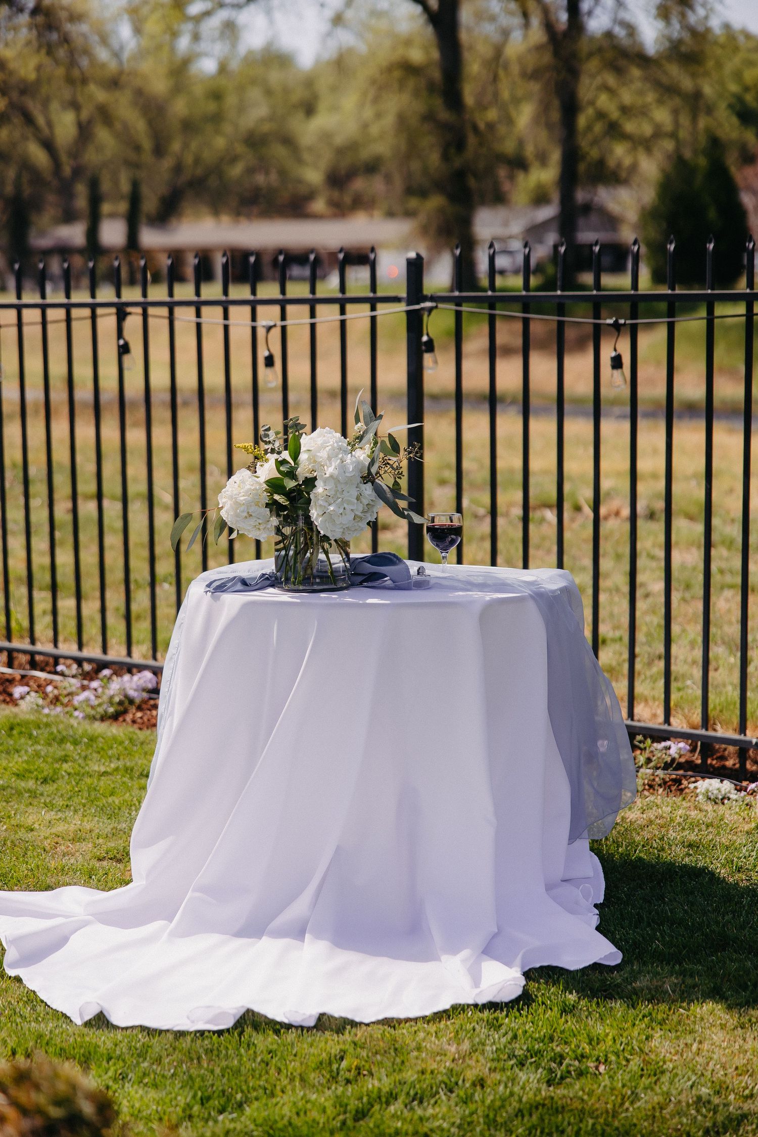 White clothed round table with floral arrangement against black iron fence outdoors.