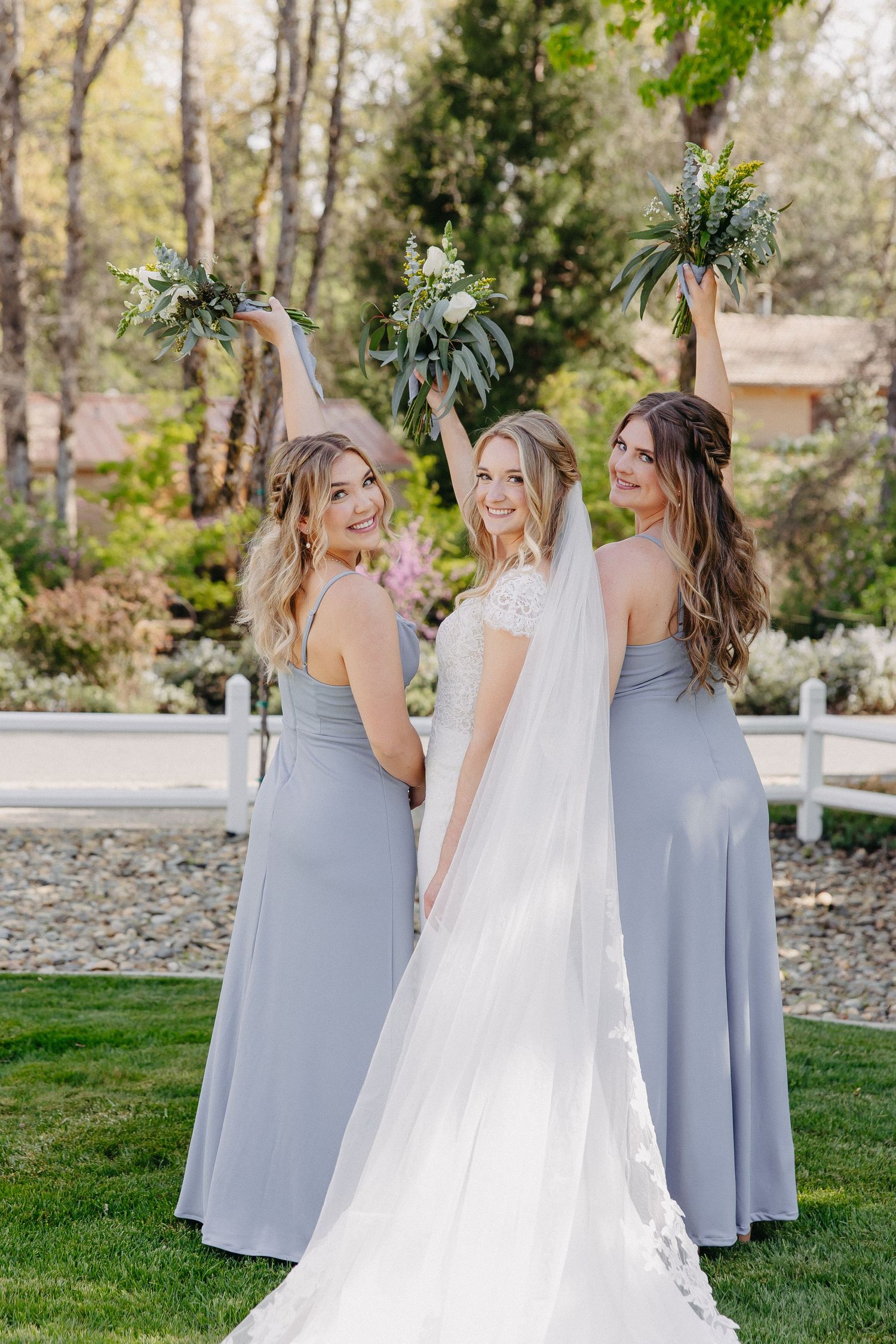 Three women in formal attire hold bouquets while posing together in a garden setting.