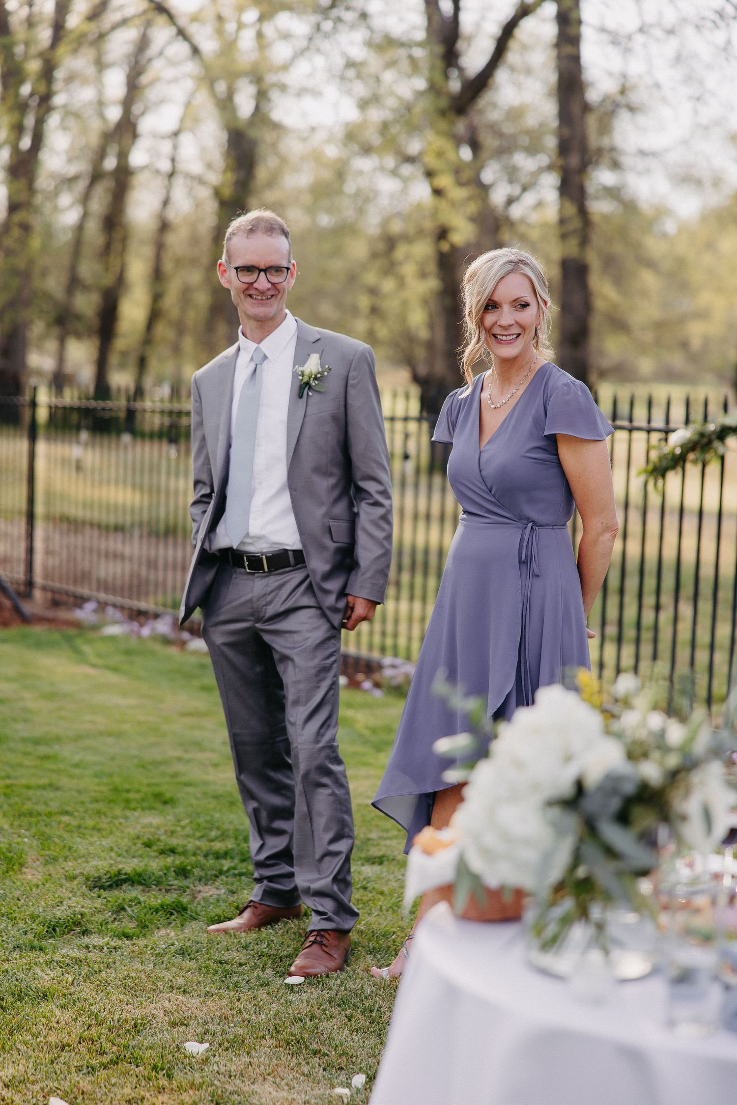Wedding guests in formal attire standing by decorated table in outdoor garden setting.