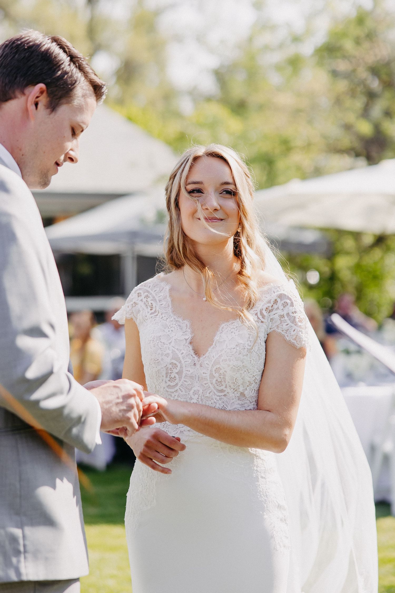 Couple exchanging rings during outdoor wedding ceremony on sunny day with white parasols in background.