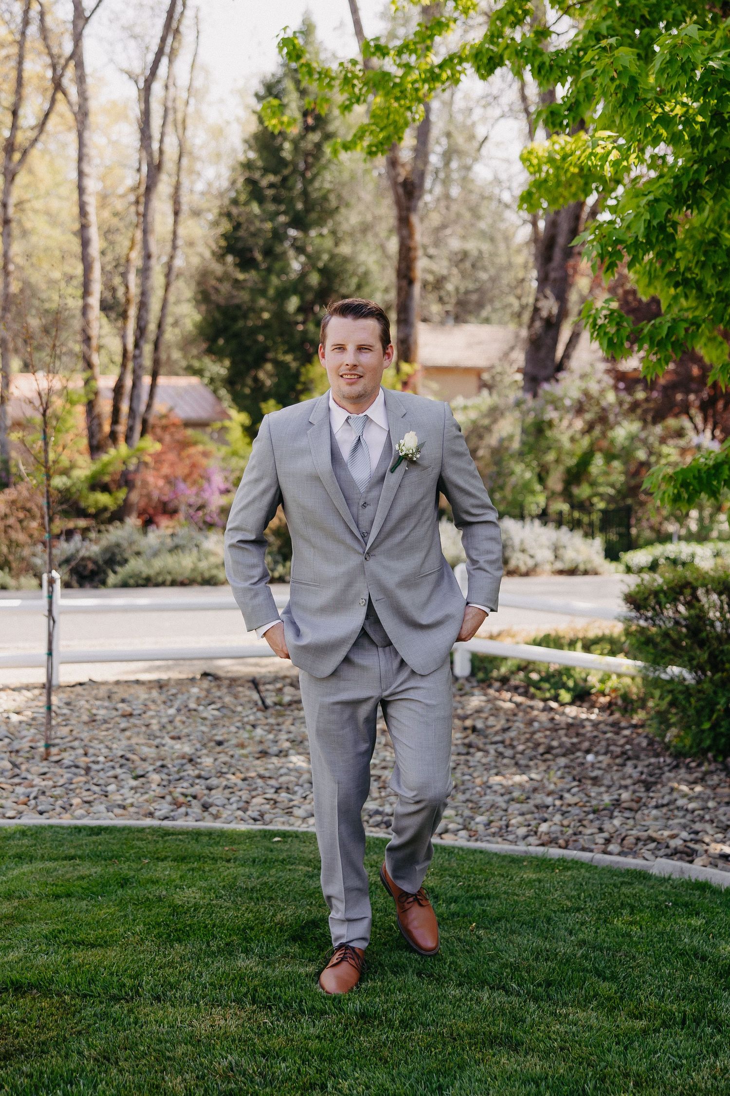 Wedding attire featuring a light grey three-piece suit with brown dress shoes in an outdoor garden setting.