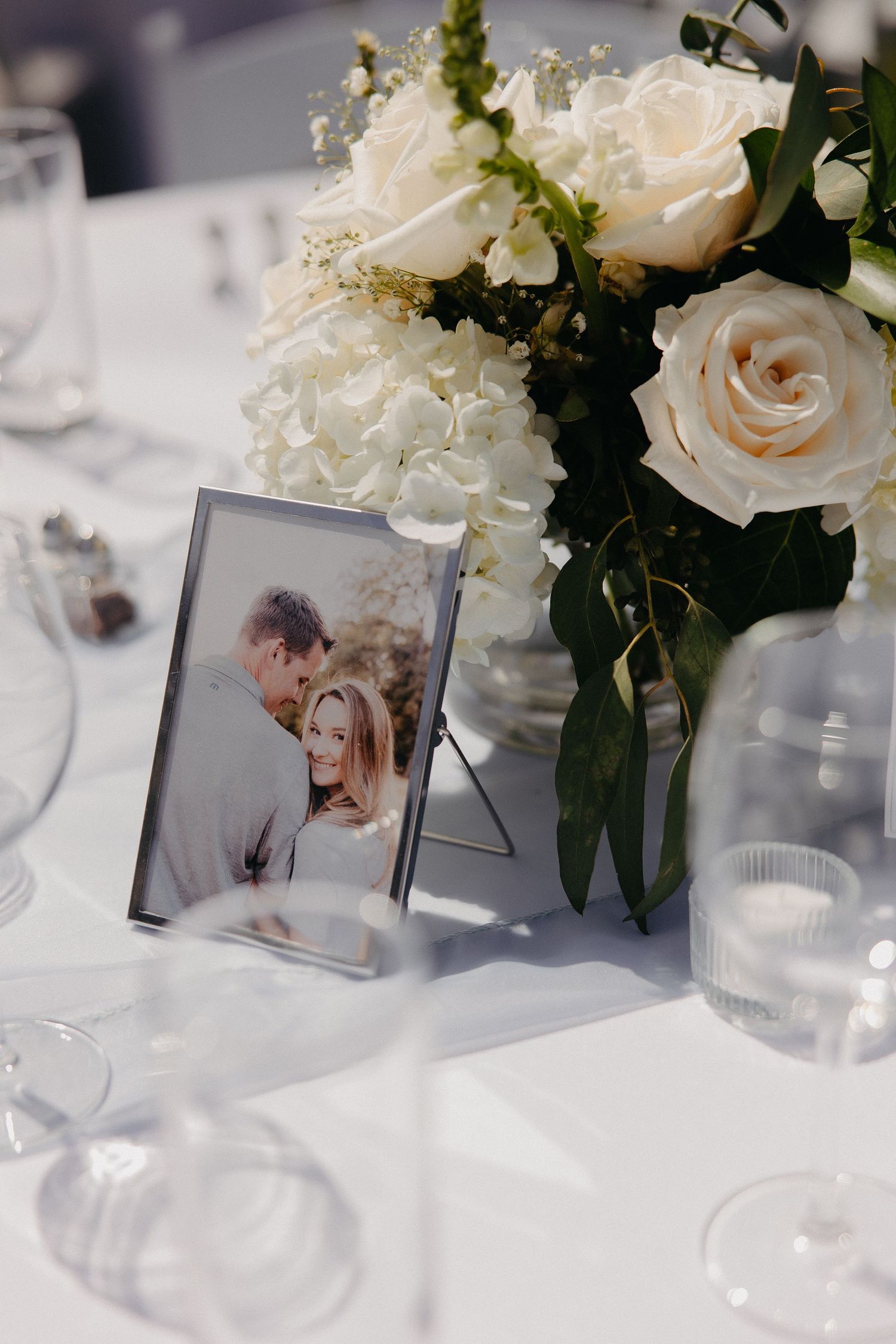 Table centerpiece with white roses and framed photograph next to elegant place settings.