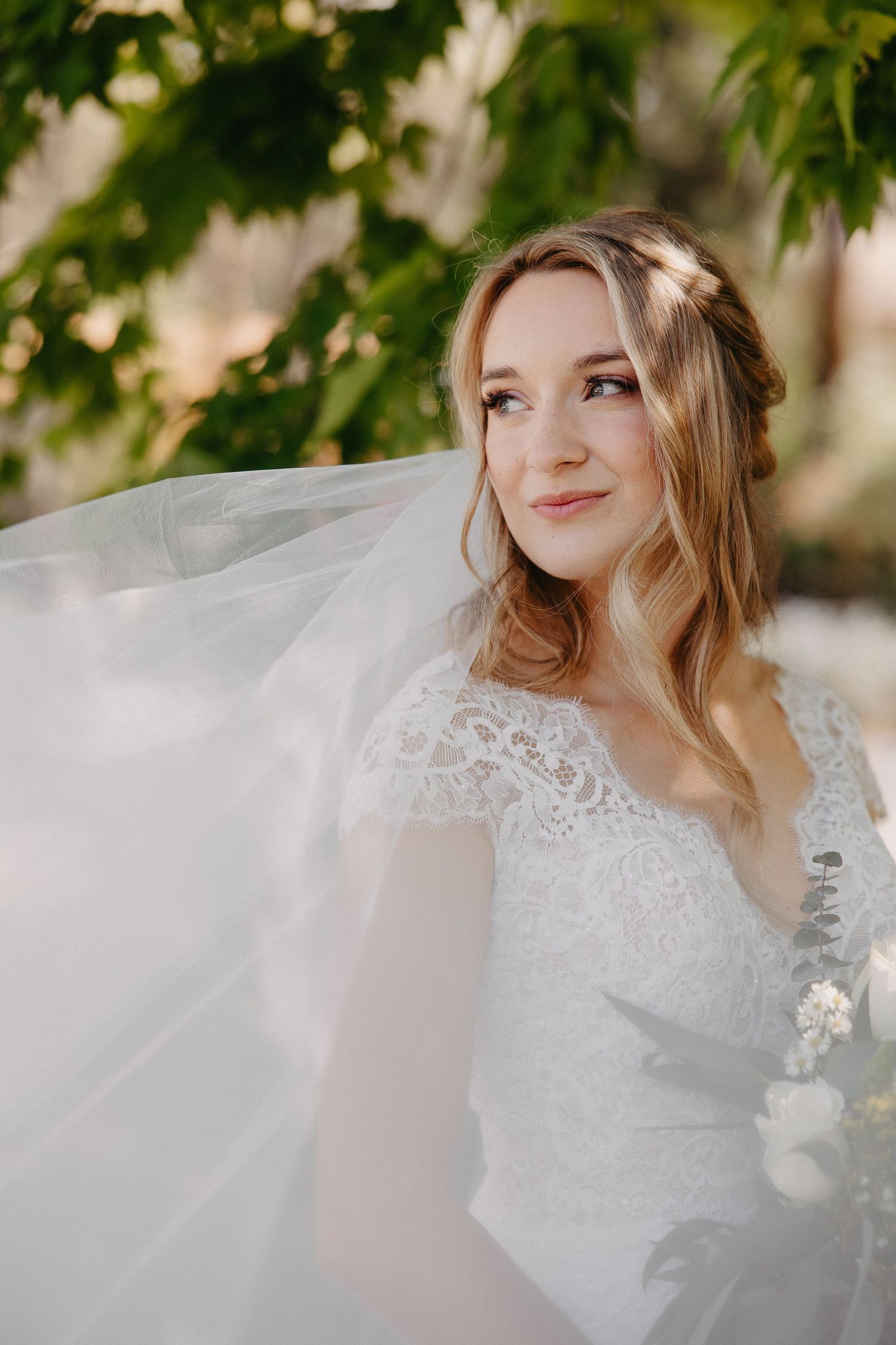 Romantic bridal portrait with flowing veil captured in soft natural lighting under green foliage.