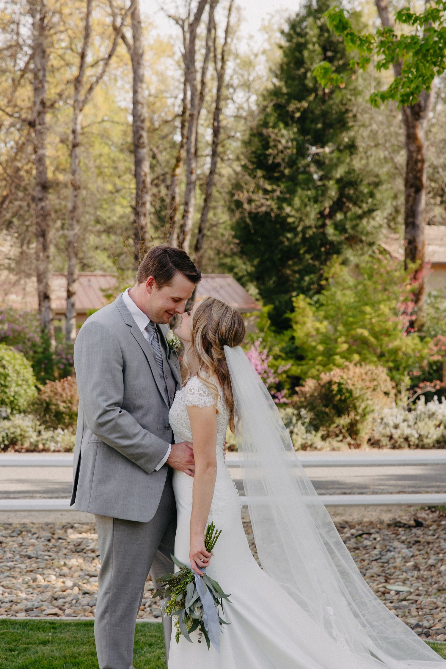 A couple shares an intimate moment in their wedding attire against a backdrop of spring foliage.