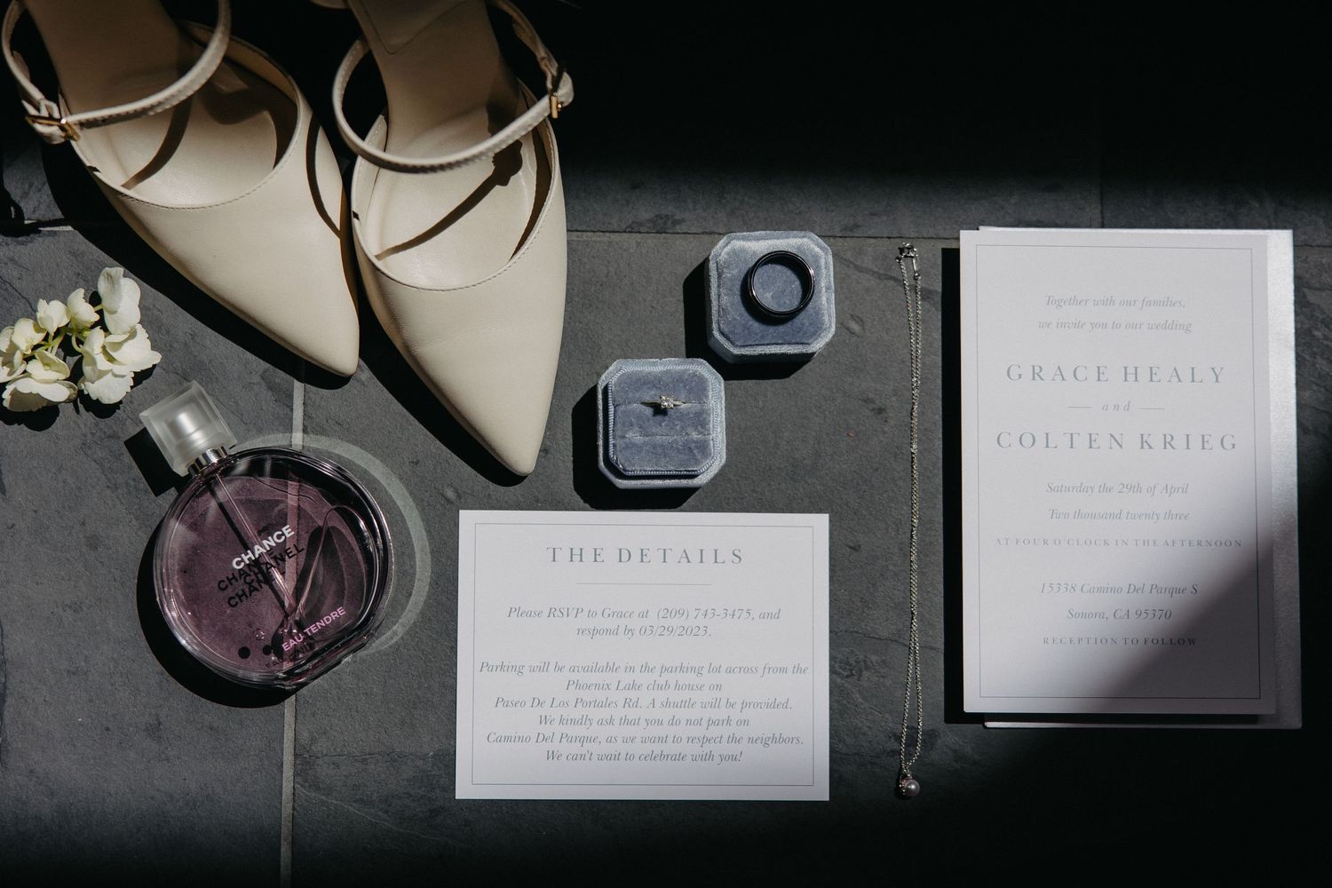 Flat lay of wedding stationery with nude heels, ring boxes and perfume bottle on dark surface.