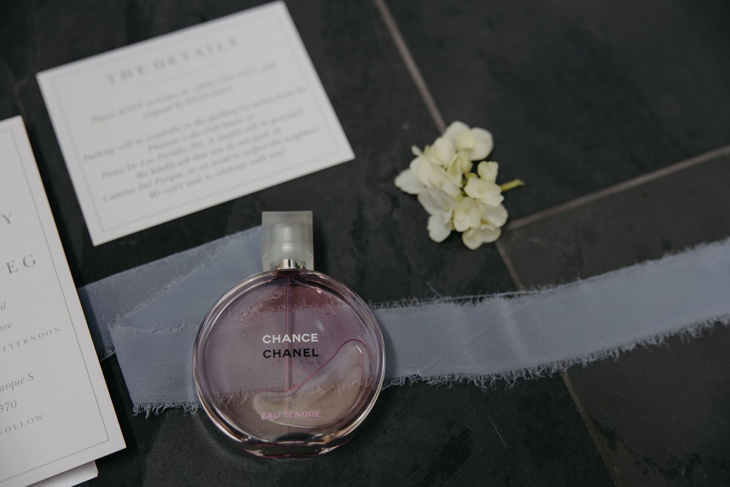 Wedding details featuring Chanel perfume bottle and invitation cards on dark surface.